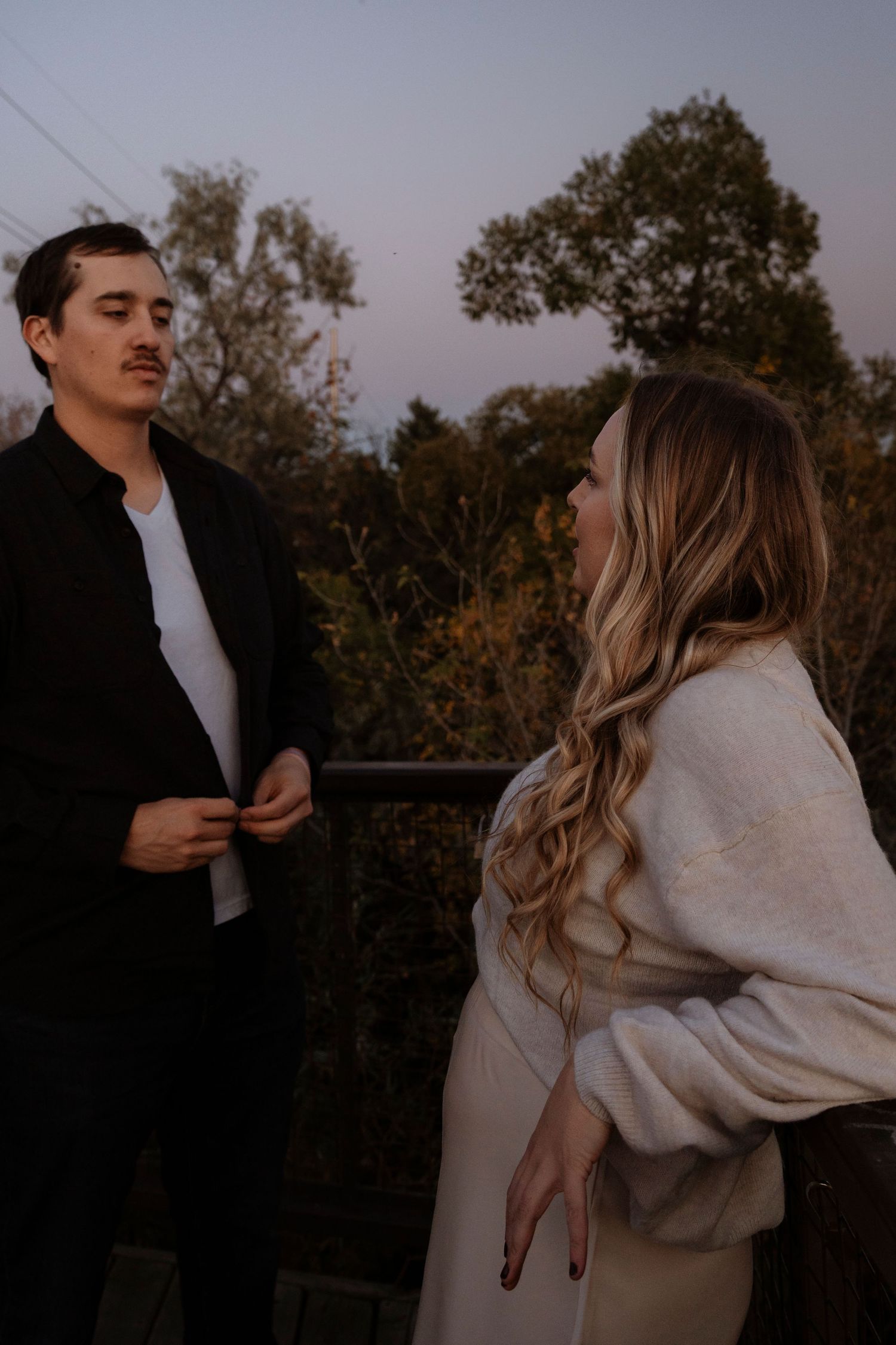 Couple standing on outdoor balcony at dusk, woman with long blonde hair in white sweater, man in black jacket.