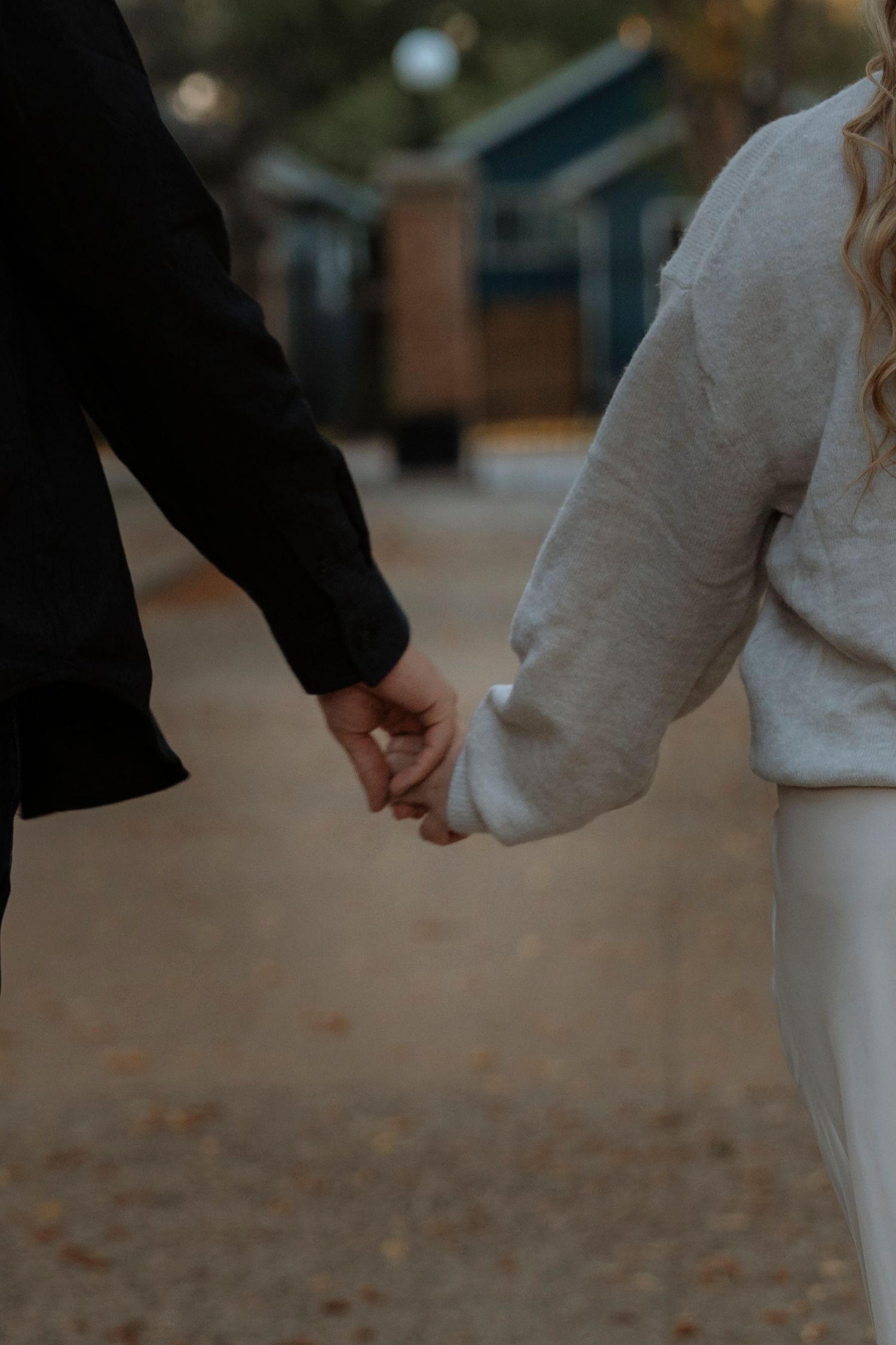 Couple holding hands while walking outdoors, one wearing black jacket and other in white sweater, with blurred buildings in background.