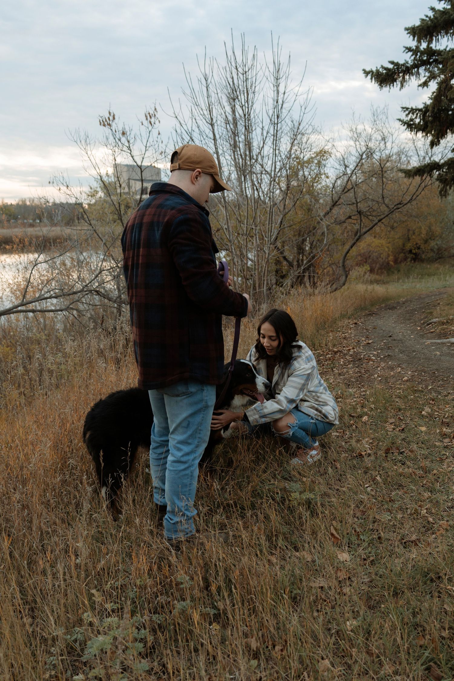 Couple with black dog on autumn trail near lake, woman crouching to pet dog while man holds leash outdoors.