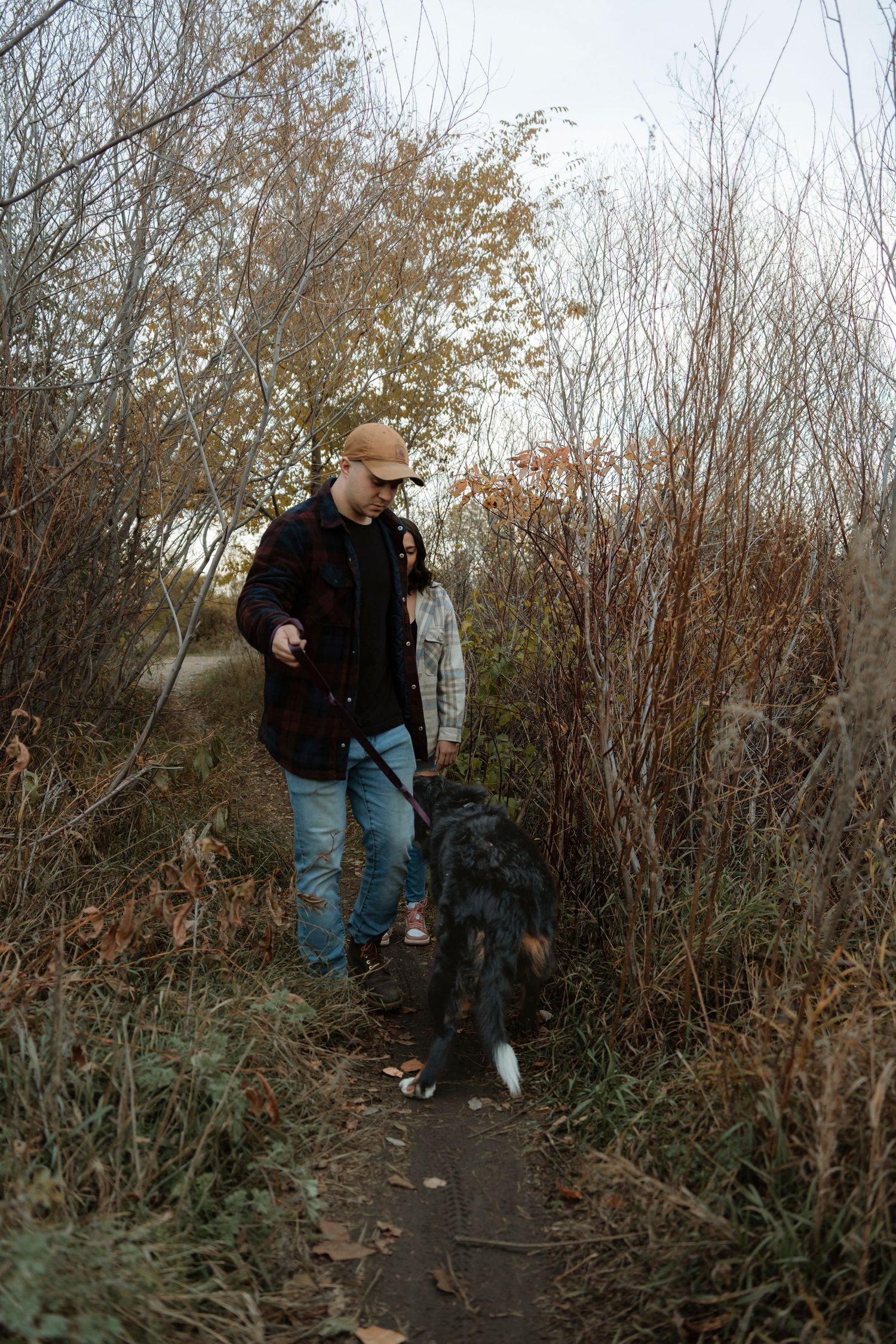 Man and woman walking a black dog on a narrow autumn trail surrounded by bare shrubs and trees.