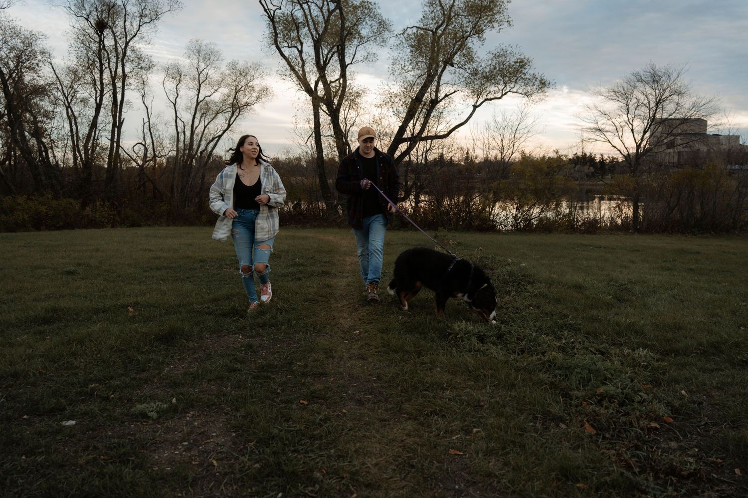 Two women walk a black and white dog in a grassy park at dusk with bare trees and a lake in the background.