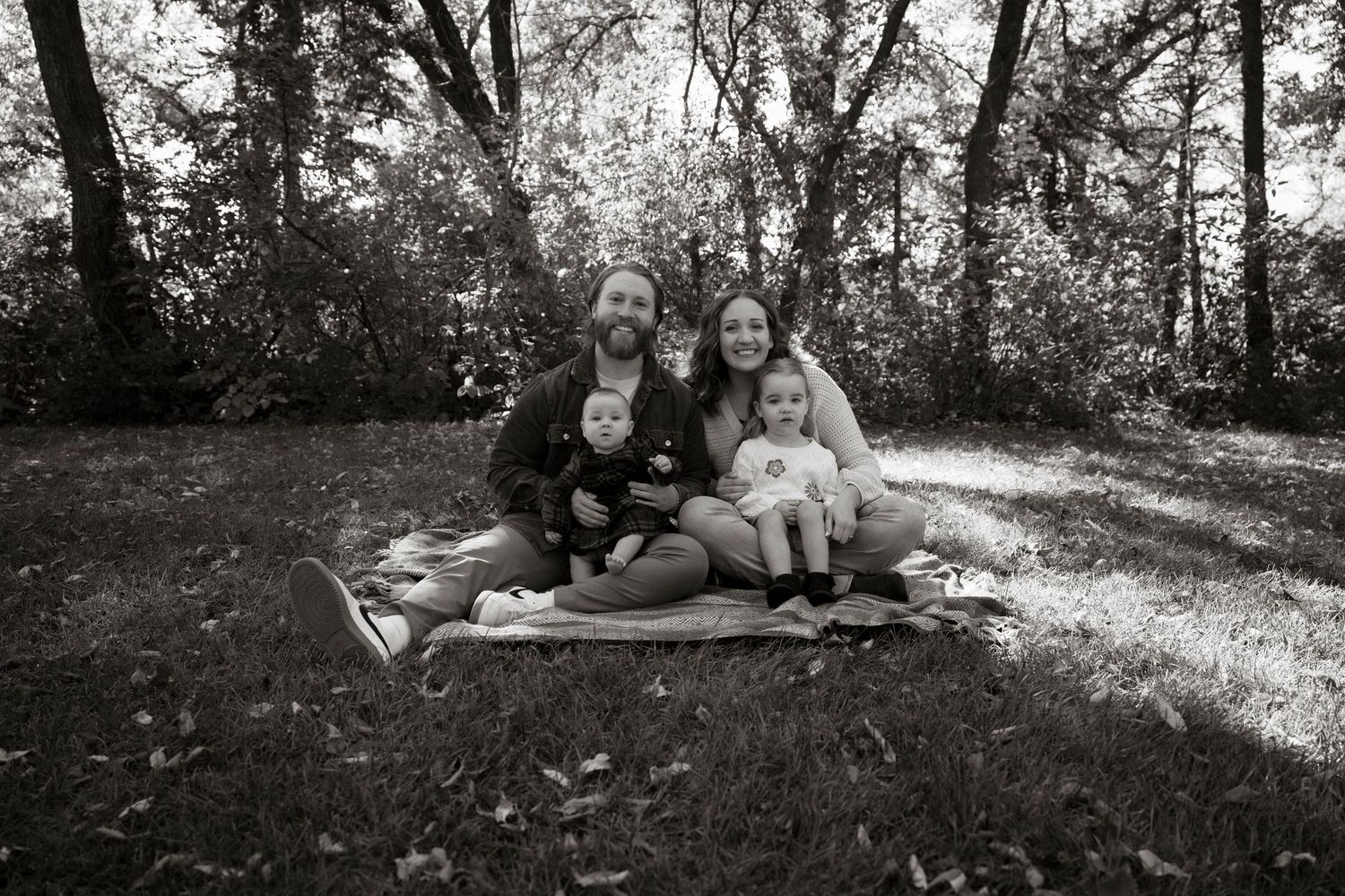 Black and white family portrait of parents with two young children sitting on a blanket in a lush wooded park.