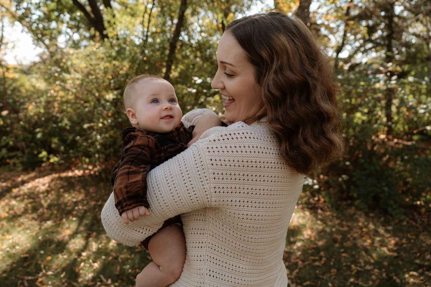 A smiling mother holds her baby in a cream knit sweater outdoors among autumn trees.