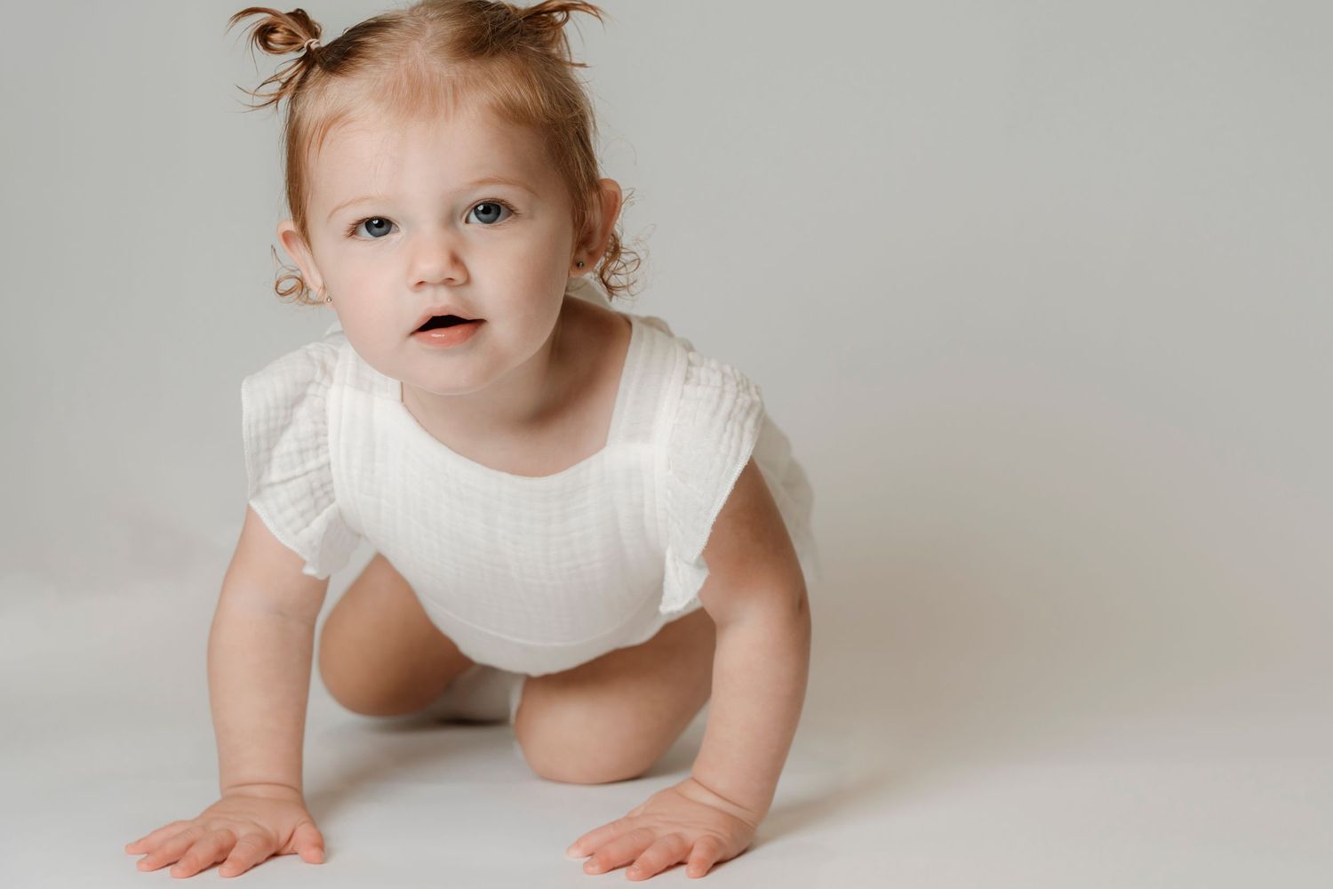 A baby girl in a white onesie crawling on a light background, looking curiously at the camera.