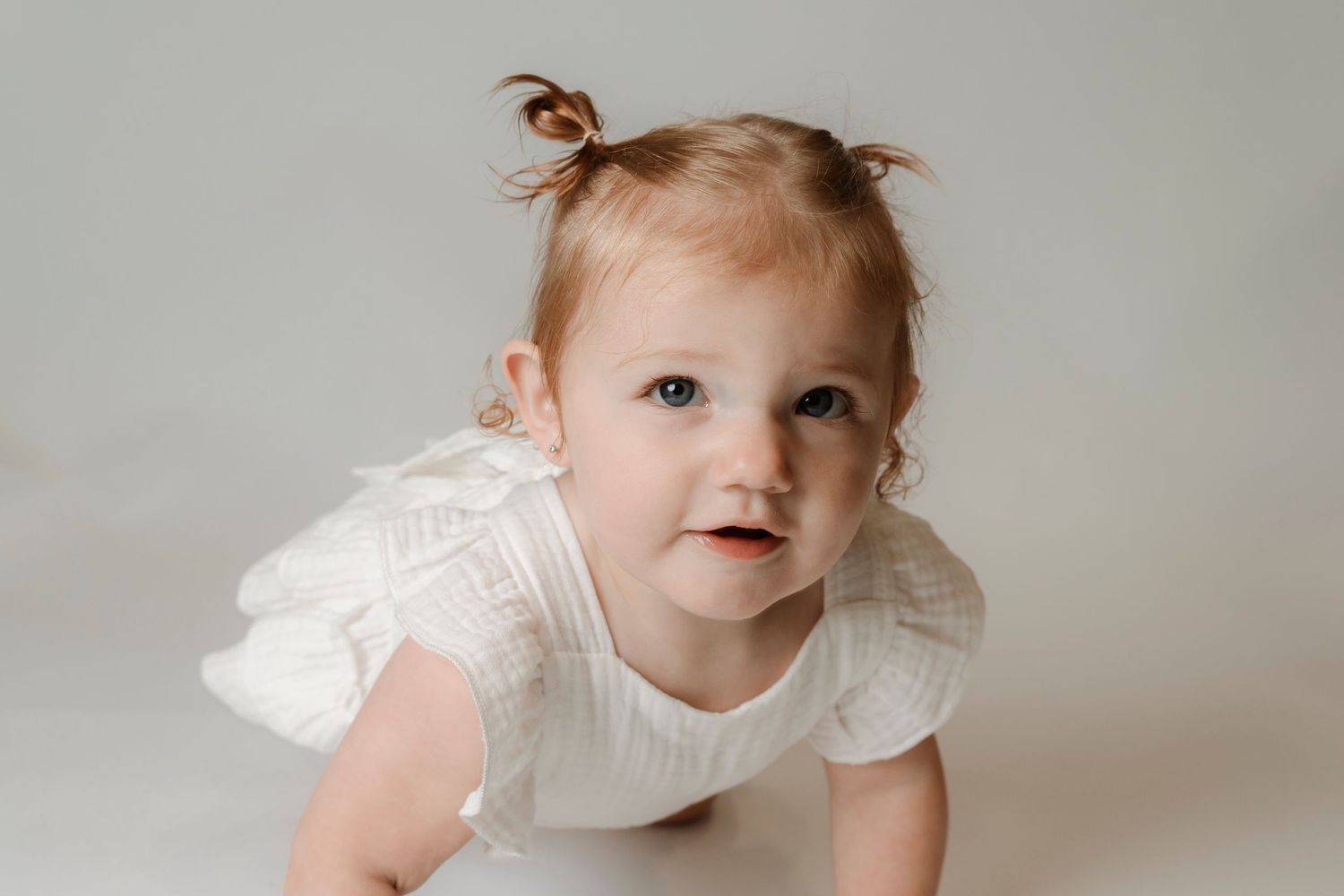 A cute baby girl with red hair in tiny pigtails wearing a white ruffle dress, crawling and looking up curiously.
