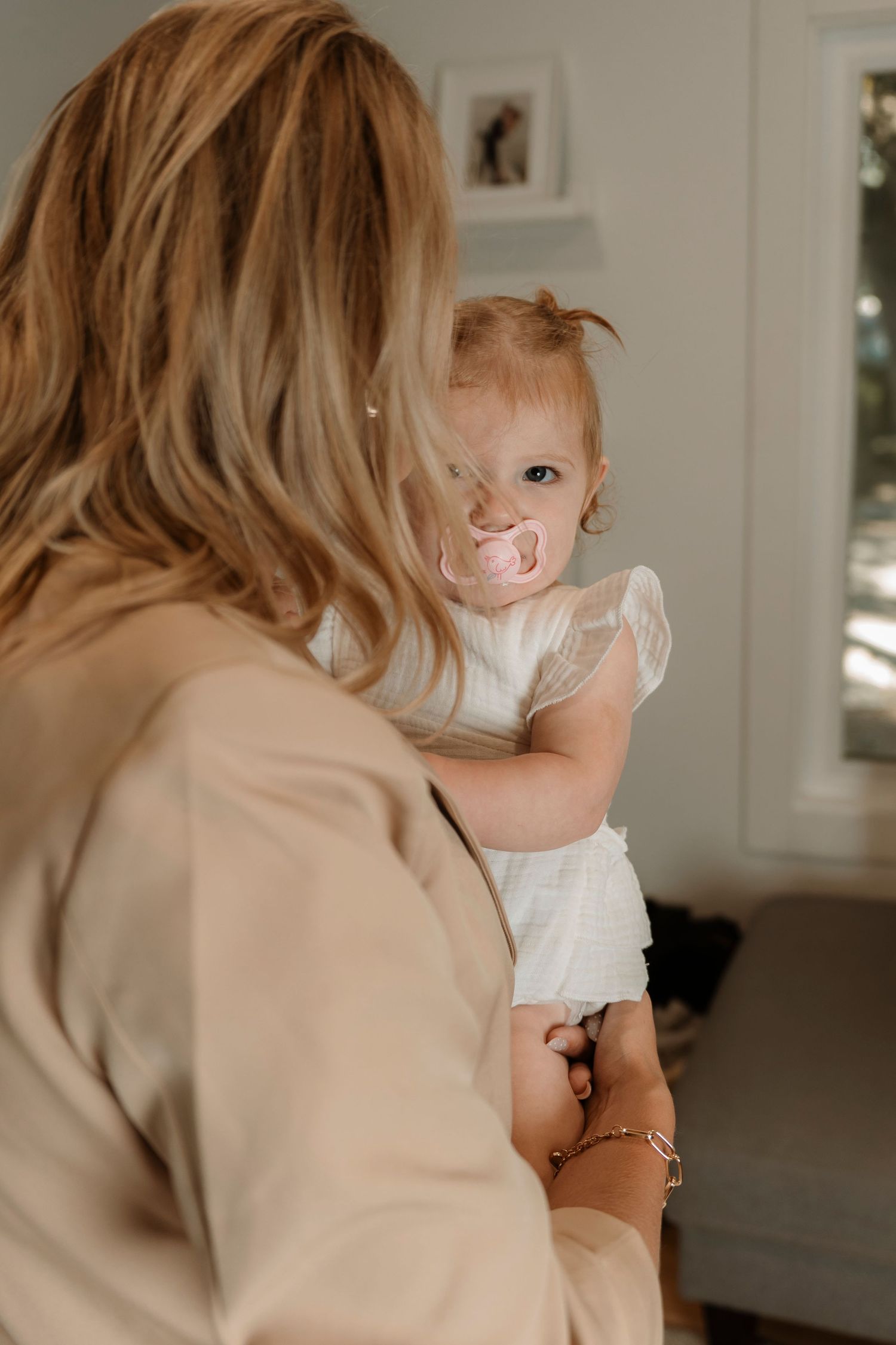 A blonde woman in a beige coat holds a baby girl in a white dress with a pink pacifier in a bright room.