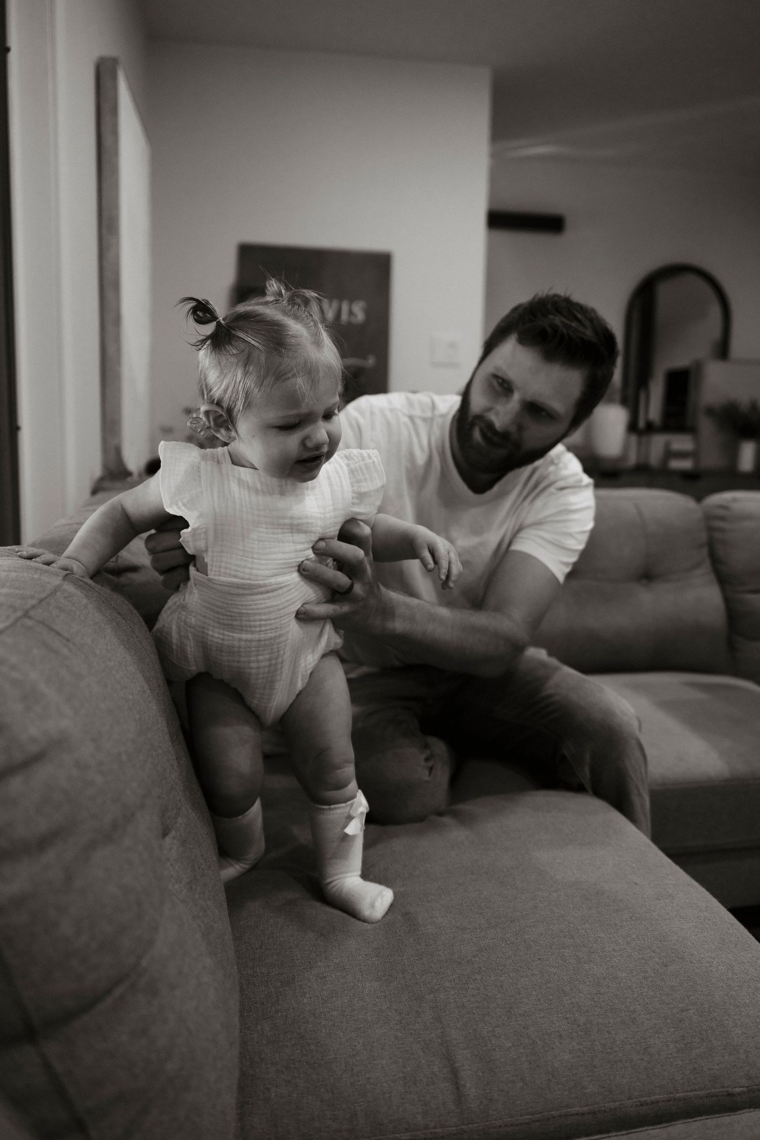 Black and white photo of a father helping his toddler daughter stand on a couch in a cozy living room.