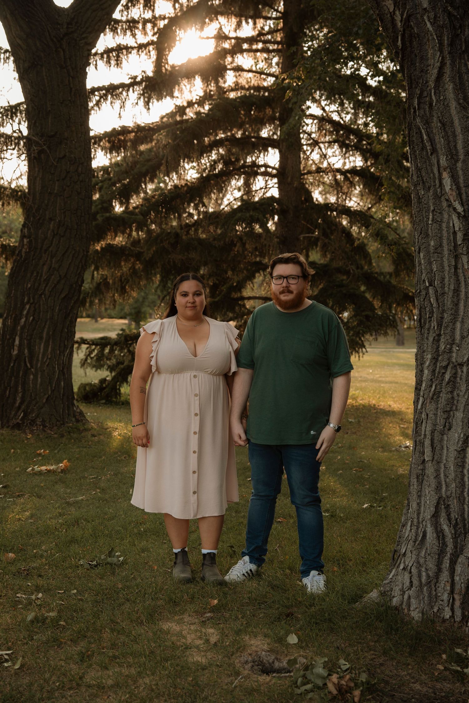 Couple standing together under large trees in a park during golden hour sunset, woman in white dress, man in green shirt.