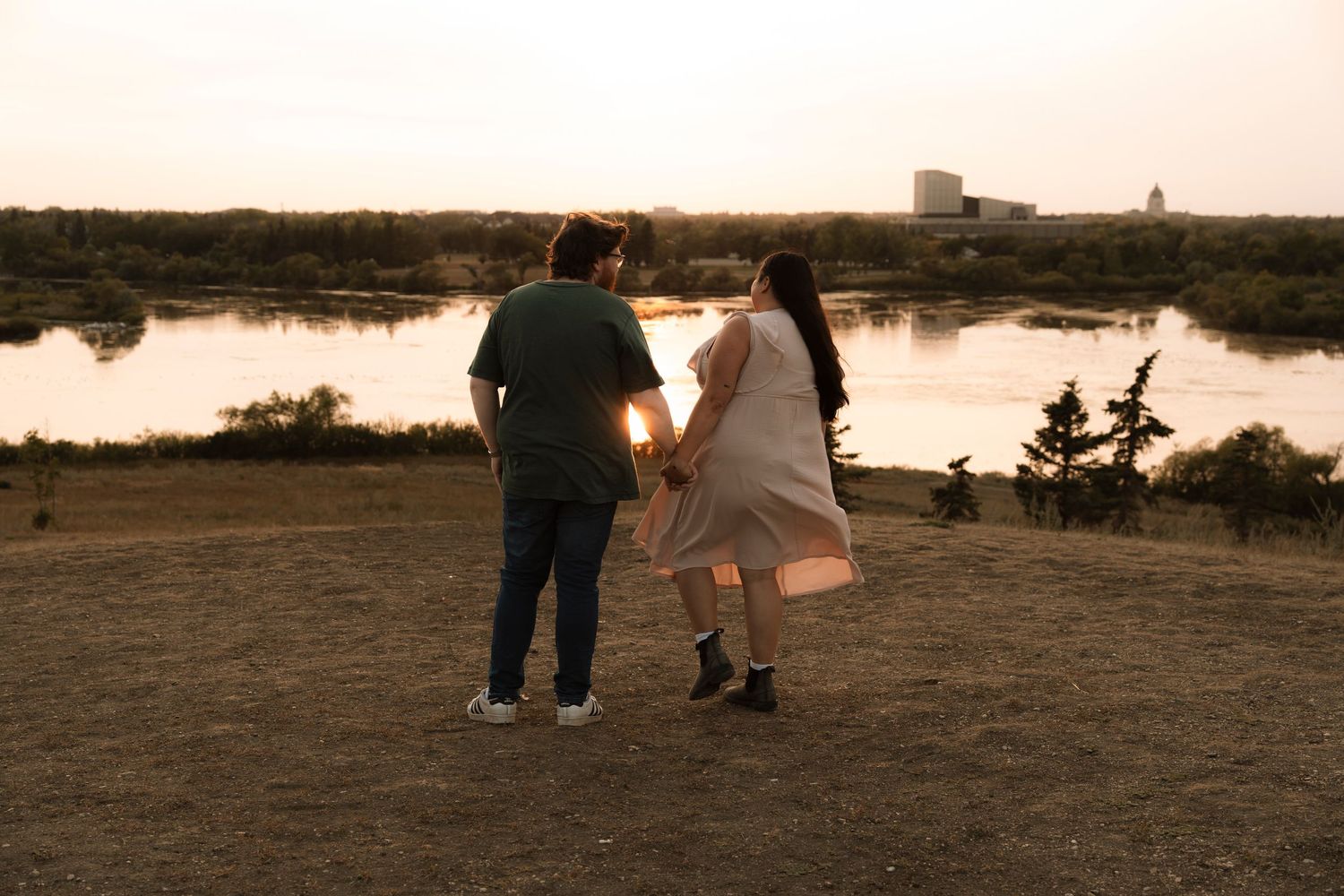 Couple holding hands at sunset by a serene lake, viewed from behind, golden hour light glowing warmly.
