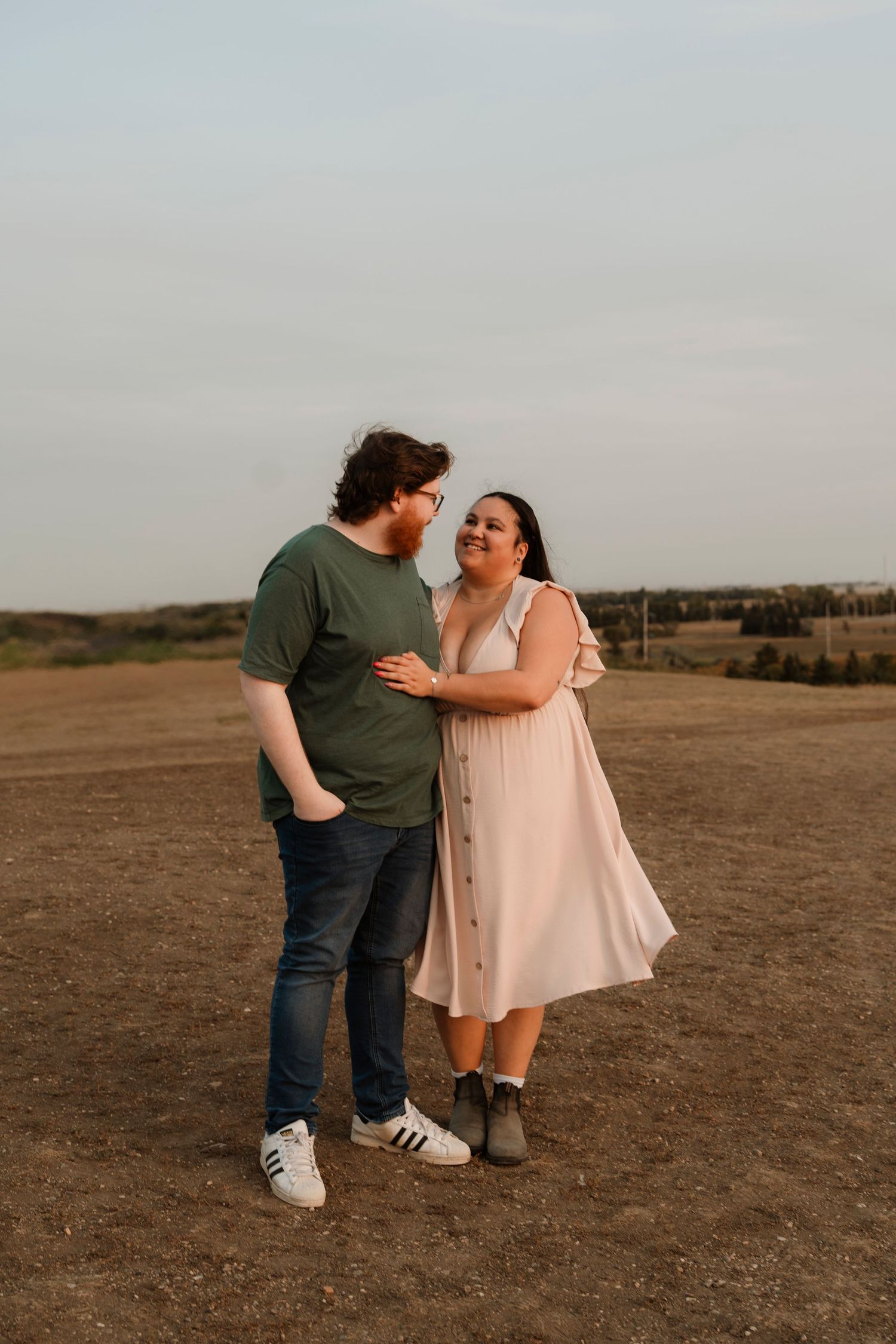 A couple smiles at each other while standing in an open field at golden hour, she in a pink dress, he in a green tee.