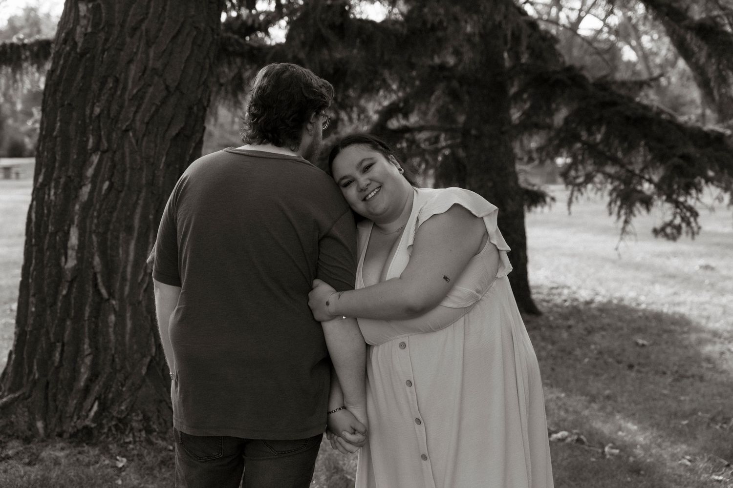 Black and white photo of a couple embracing near a large tree in a park, woman smiling warmly.