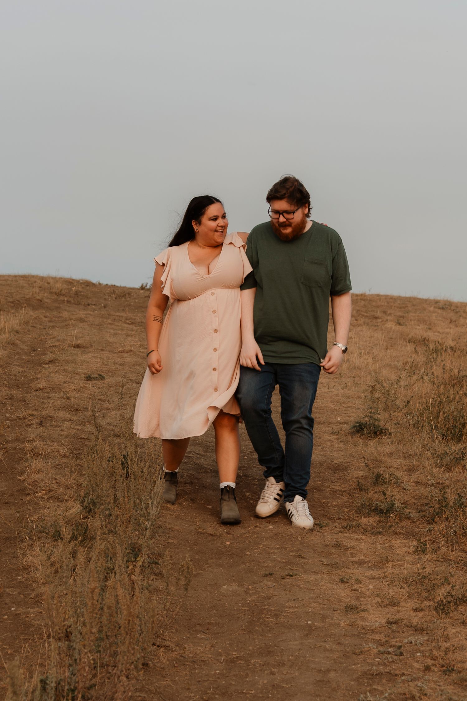 Couple walking hand in hand on a dry hillside trail, woman in pink dress and man in green shirt and jeans.