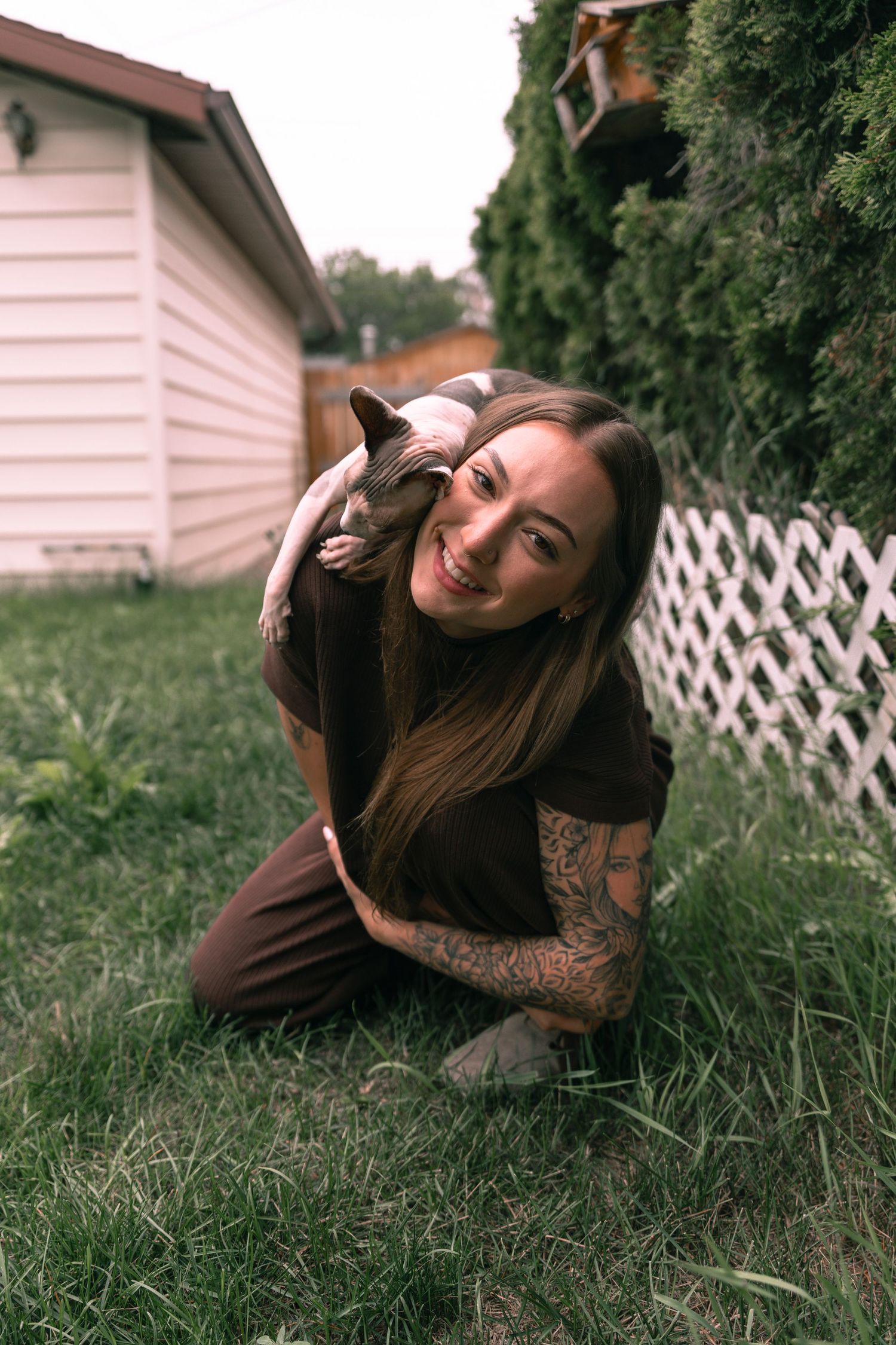 Smiling tattooed woman kneeling on grass in backyard, cuddling small dog against her cheek near white fence.