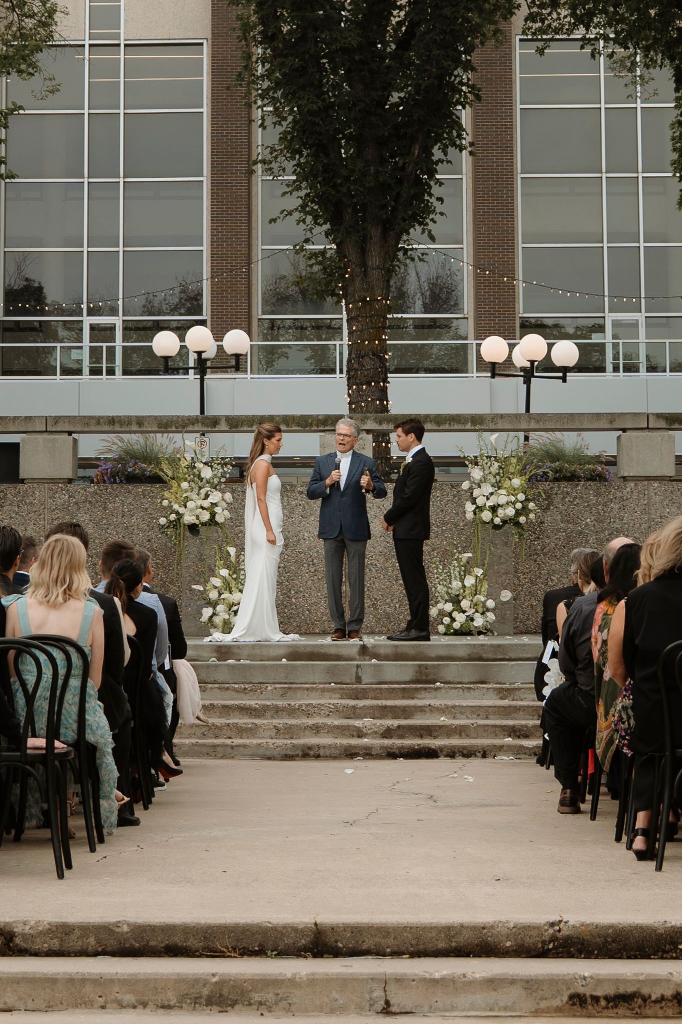 Outdoor wedding ceremony on steps with bride, groom, and officiant, flanked by white floral arrangements and seated guests.