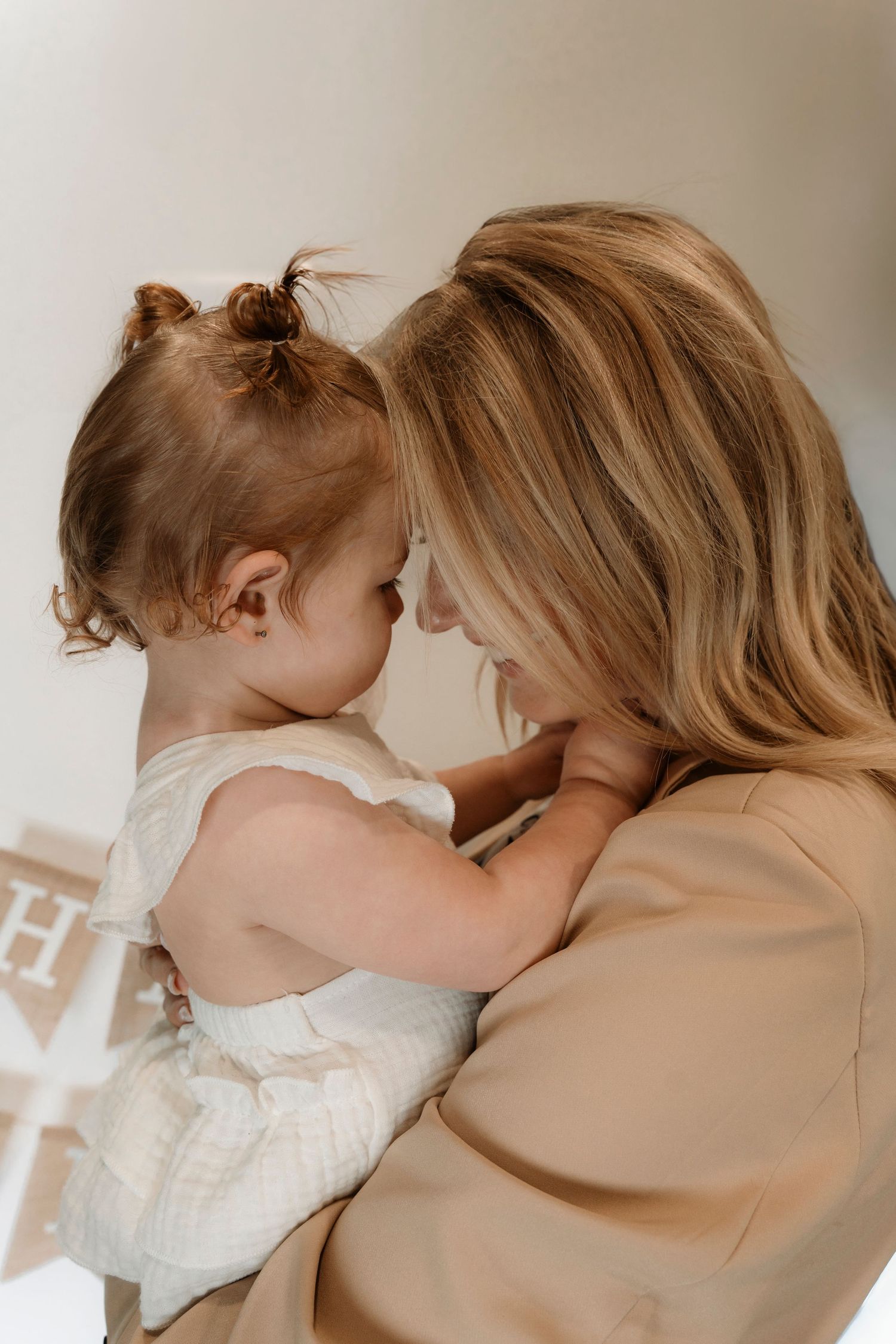A blonde mother tenderly holds her toddler daughter close, sharing a gentle forehead-to-forehead moment in soft light.