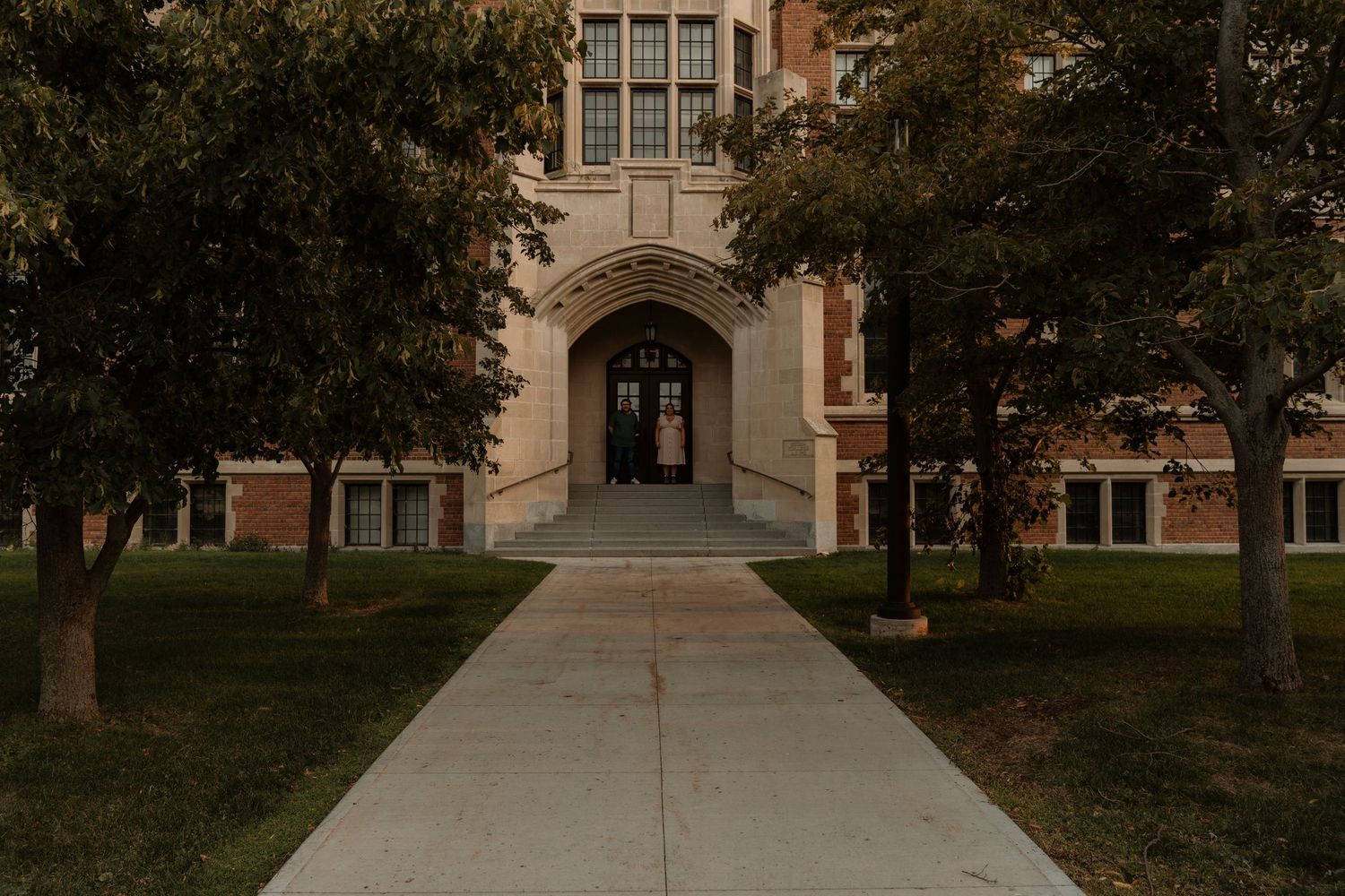Historic brick university building with arched entrance, flanked by mature trees lining a concrete walkway.