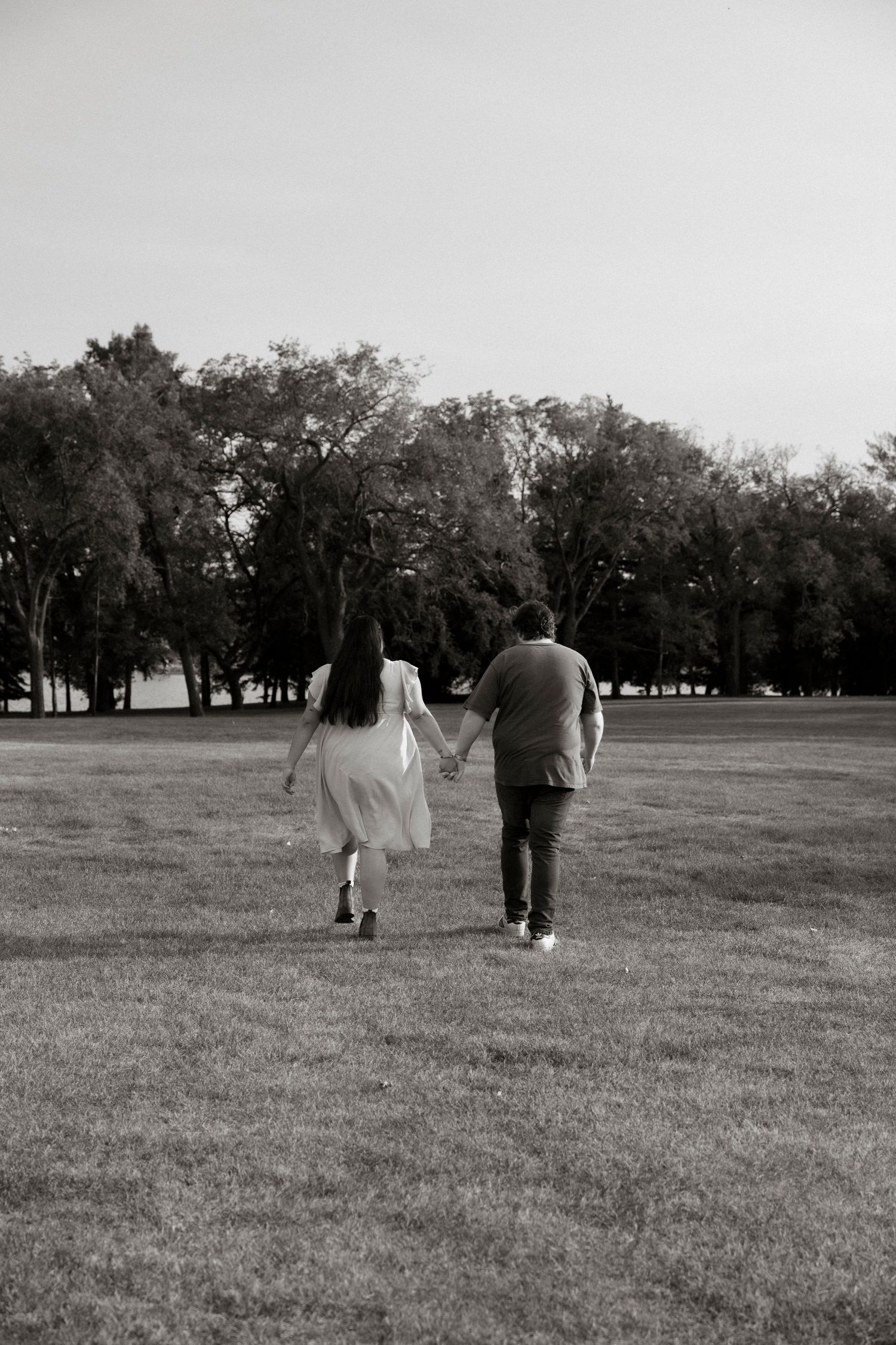 Black and white photo of a couple holding hands walking across a grassy park with trees in the background.