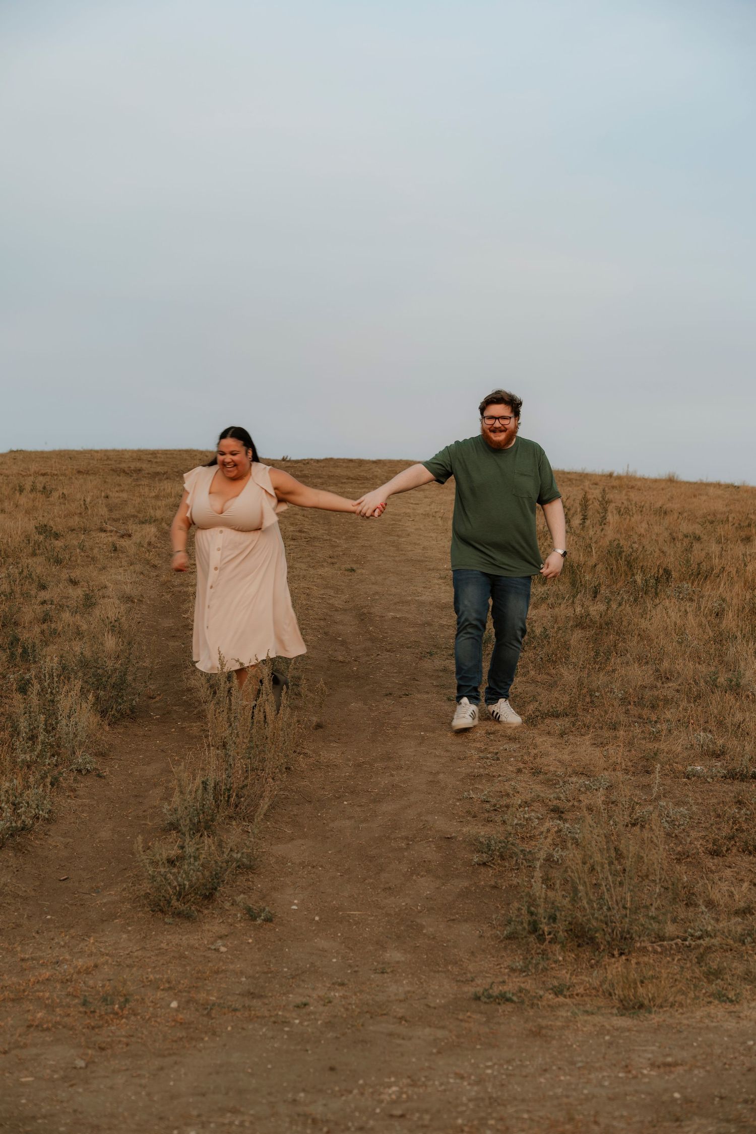 A couple holds hands while walking on a dirt path through a golden field, woman in pink dress and man in green shirt.