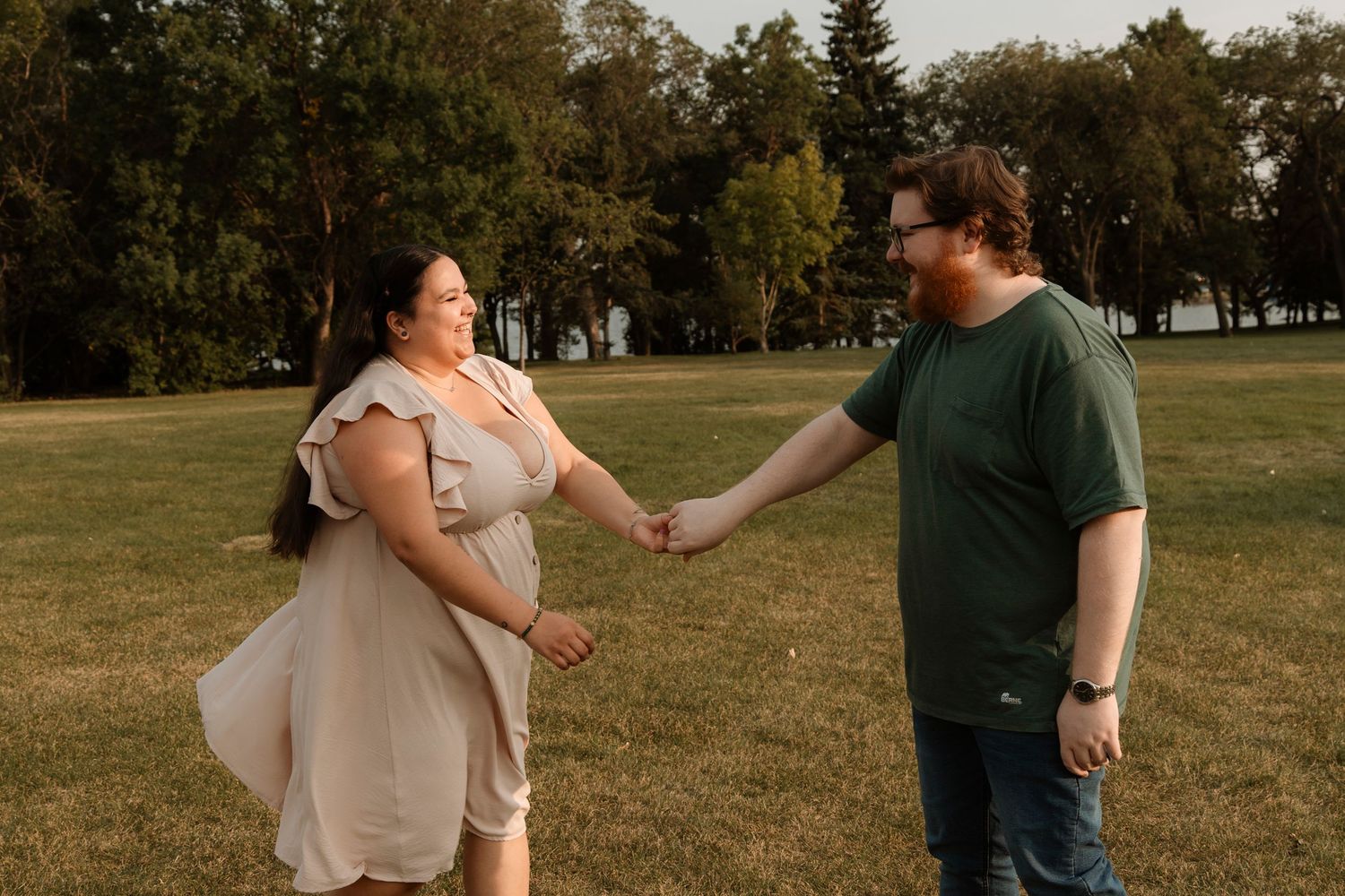 Couple holding hands and smiling in a green park, woman in white dress and man in green shirt, golden hour outdoor photo.