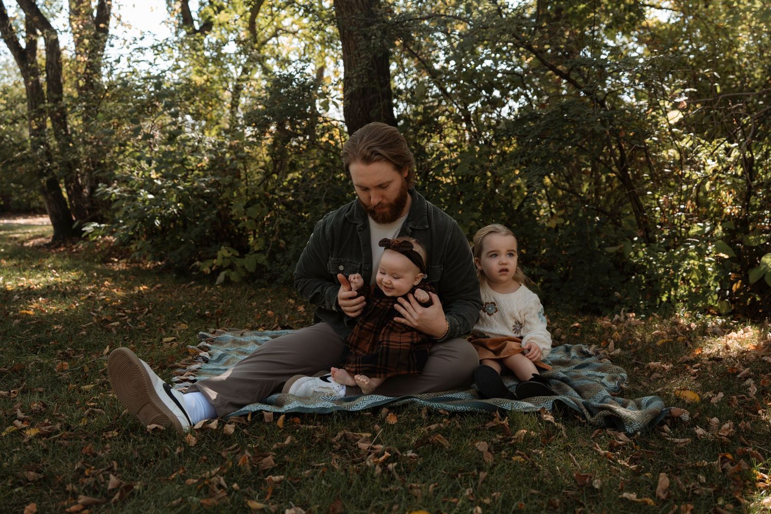 A bearded father sits on a blanket in a park with two young children, looking at a phone together.