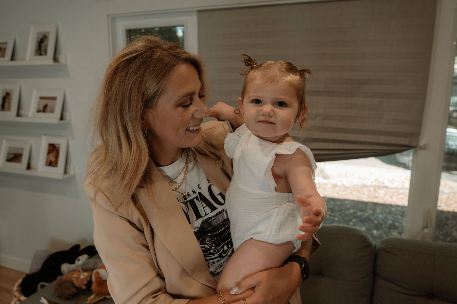 A smiling blonde woman holds a cute baby girl in a white dress in a cozy living room.