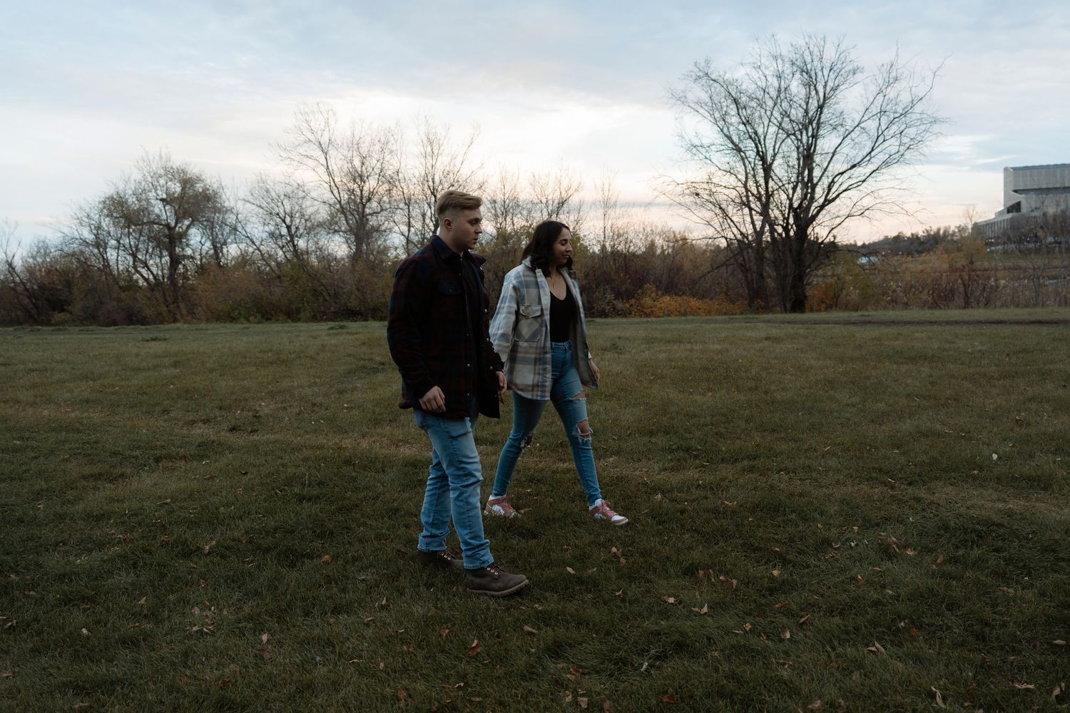 Couple walking together on green grass field at dusk with bare trees and cloudy sky in background.
