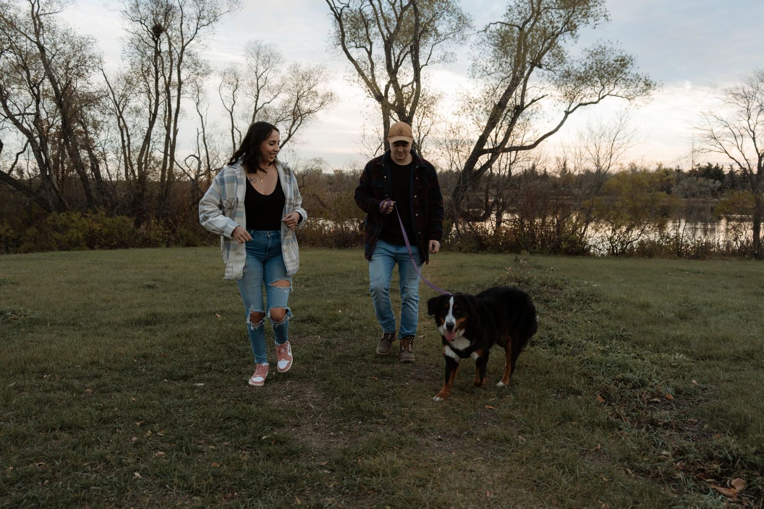 A couple walks their Bernese Mountain Dog on a grassy field near a lake surrounded by bare autumn trees.