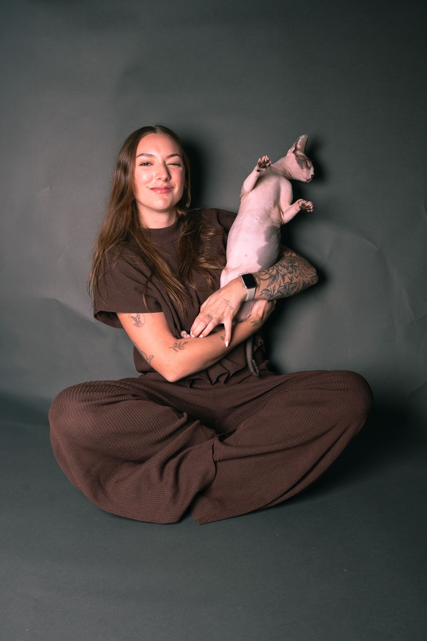 Woman in brown outfit sitting cross-legged on gray background, smiling while holding a hairless Sphynx cat up.