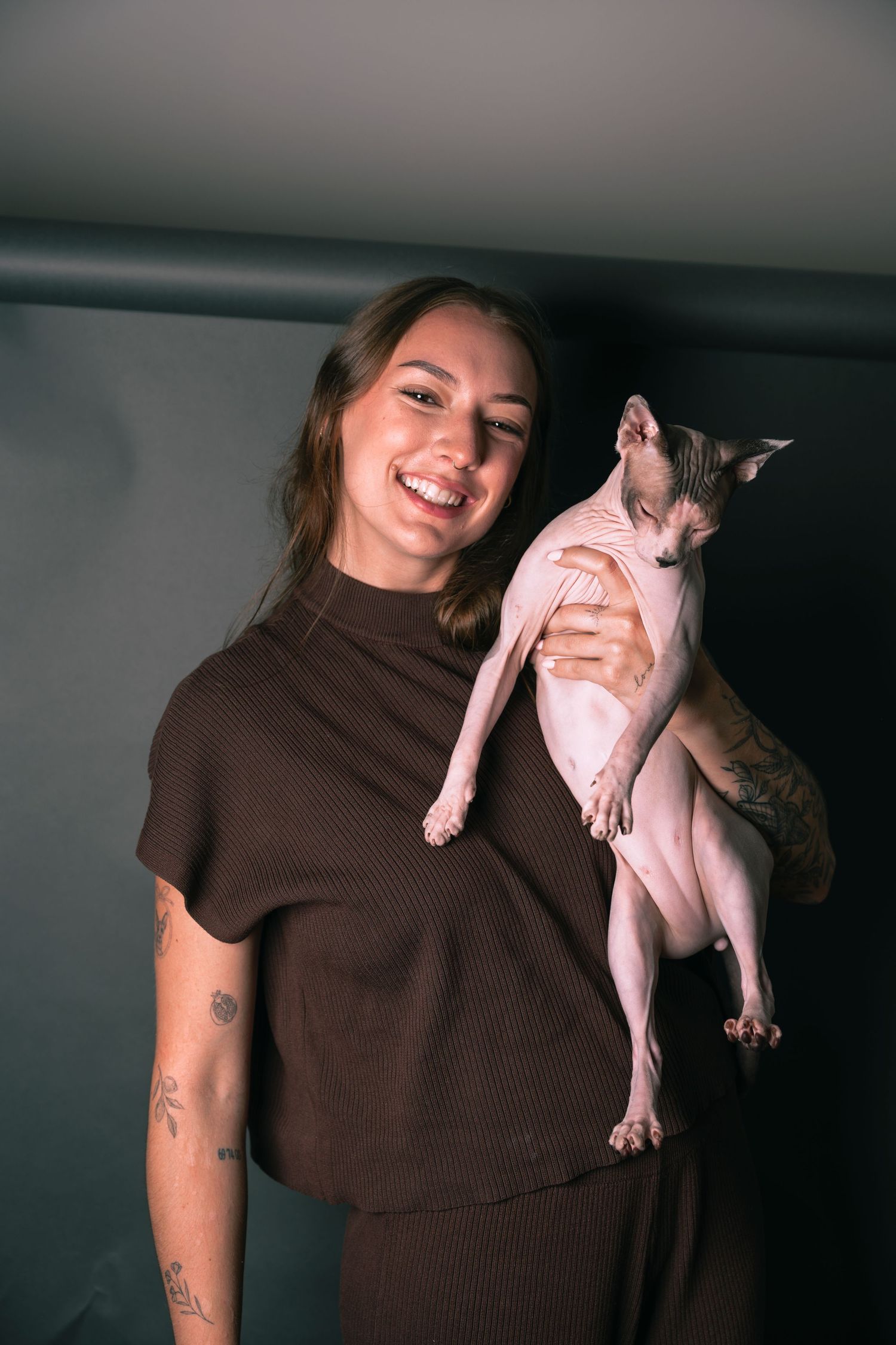 Smiling young woman with tattoos holding a hairless Sphynx cat on her shoulder against a dark background.