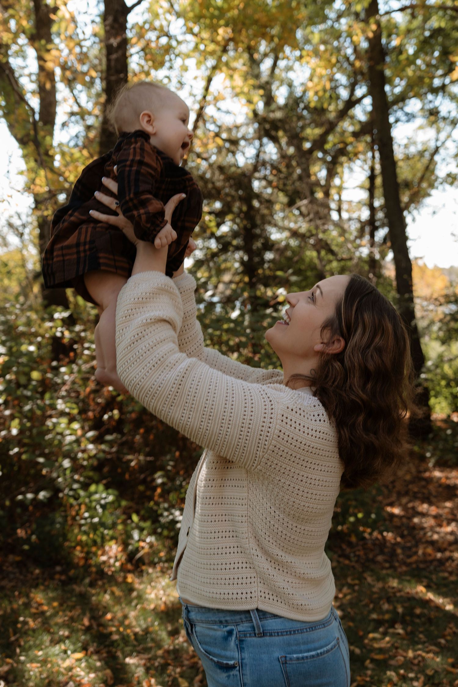 A mother lifts her smiling baby in the air outdoors among autumn trees, both wearing cozy fall outfits.