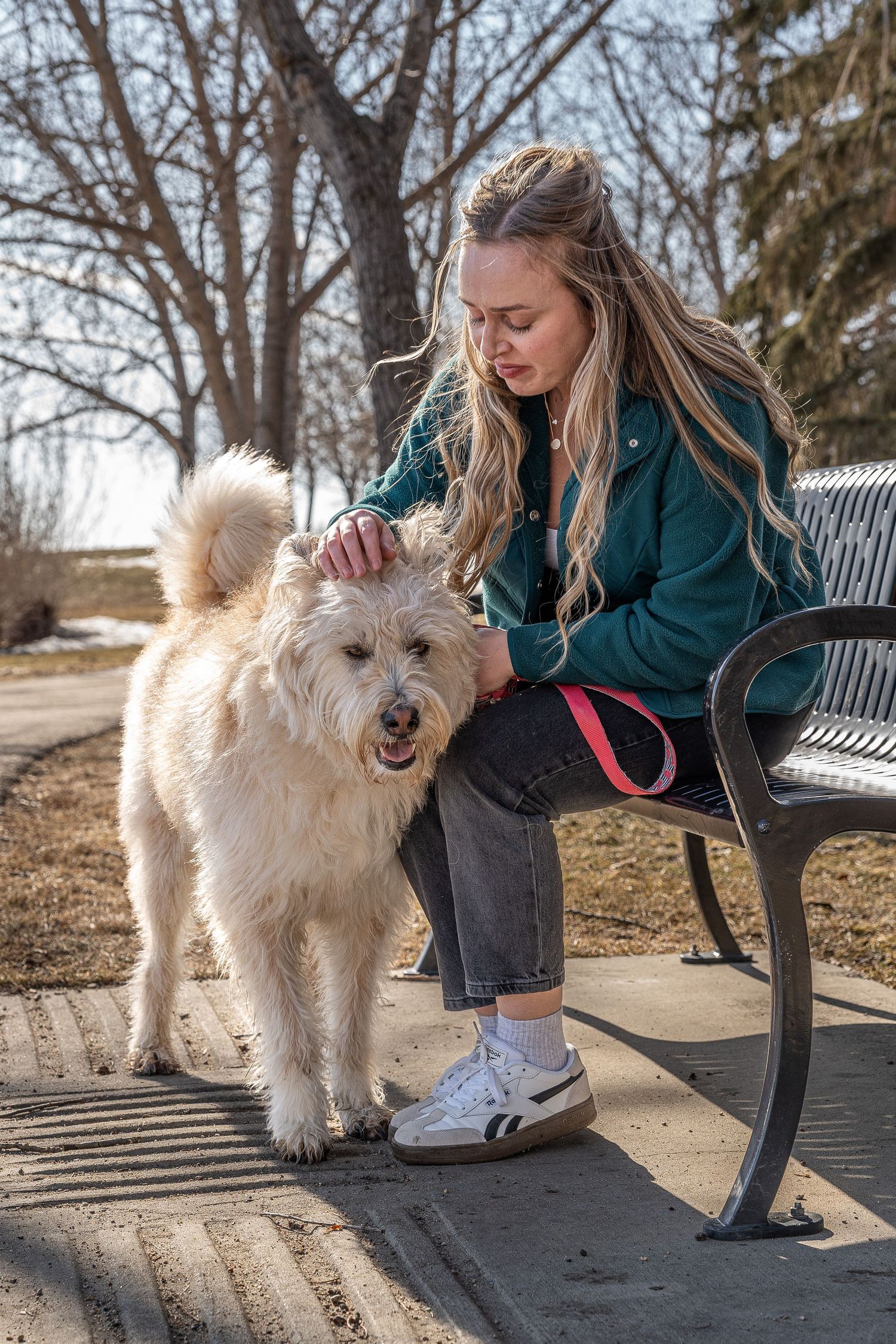Young blonde woman in teal jacket petting a fluffy white dog on a park bench outdoors. Saskatchewan Photographer.
