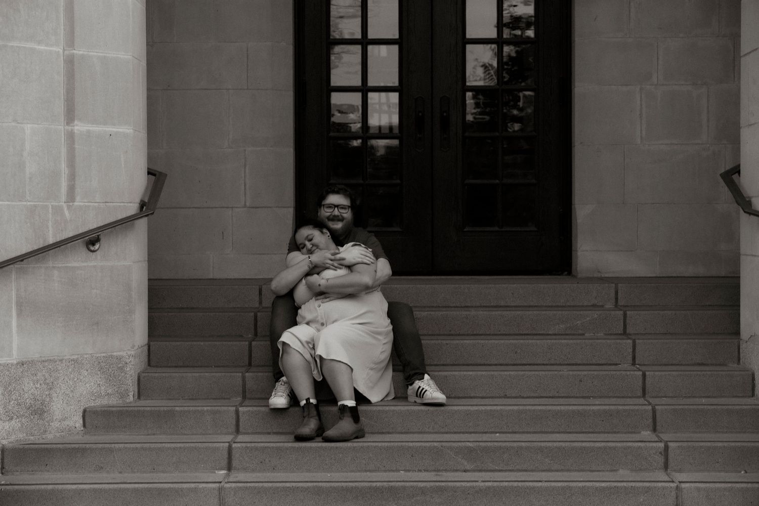 Black and white photo of a person sitting on stone steps holding a baby outside a building with large doors.