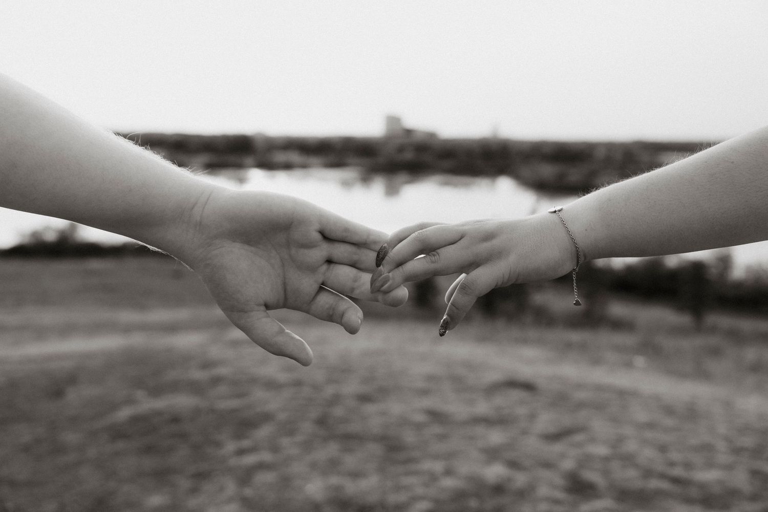Black and white photo of two hands gently touching fingertips outdoors near water, symbolizing connection and love.