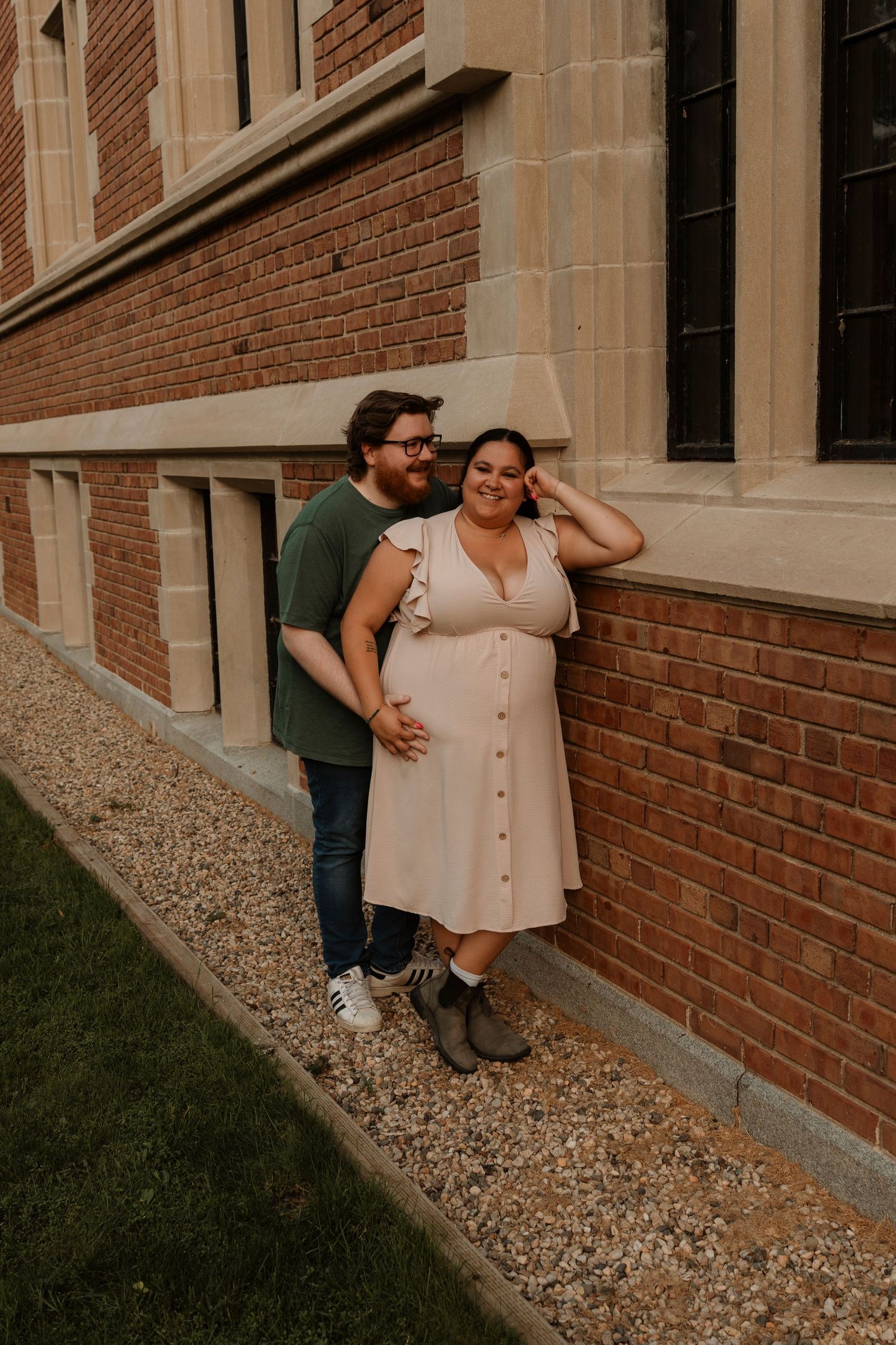 Couple posing against brick building wall, woman in pink dress with man in green shirt, maternity photo session outdoors.