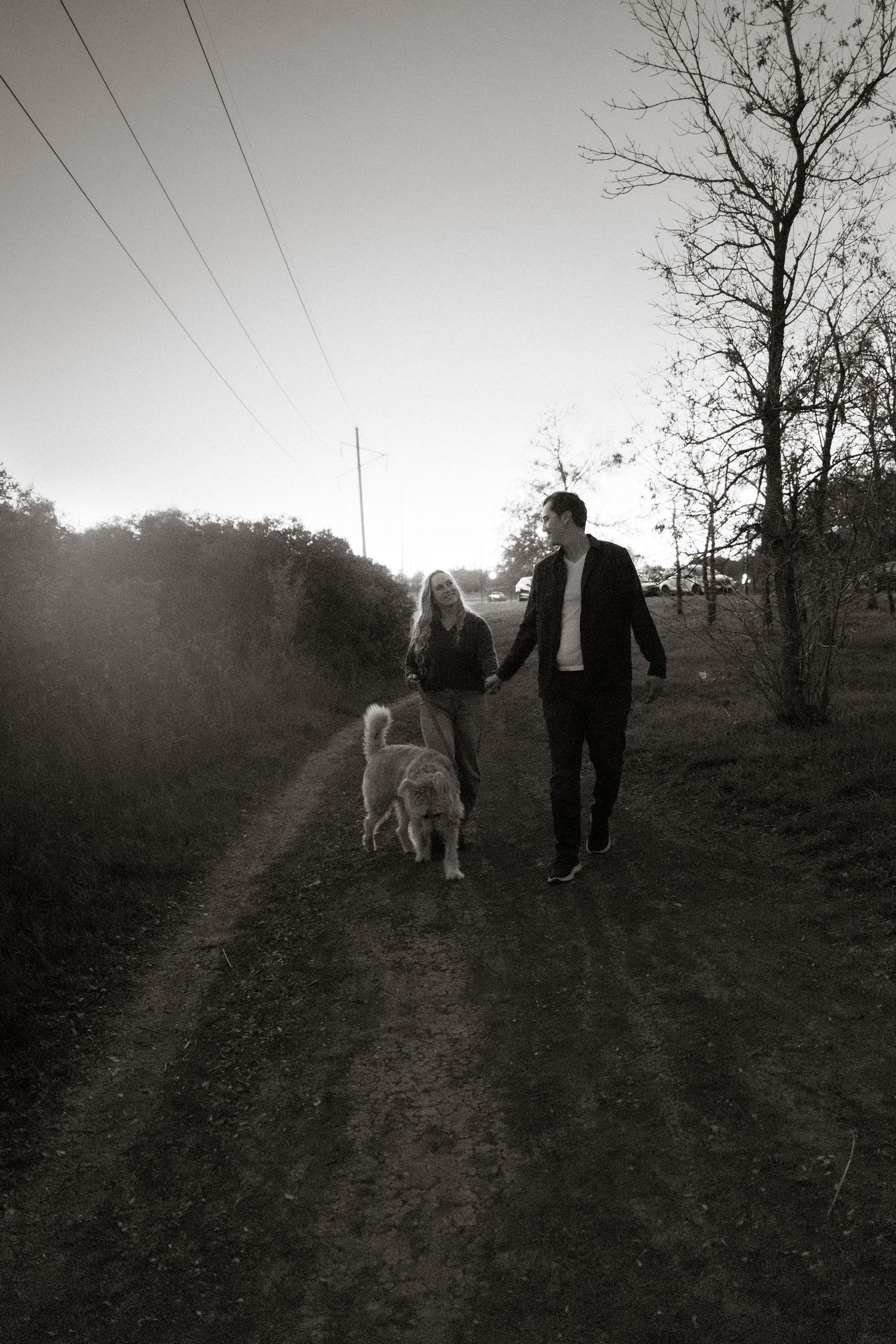 Couple holding hands walking dog on rural dirt path, black and white photo with bare trees and power lines.