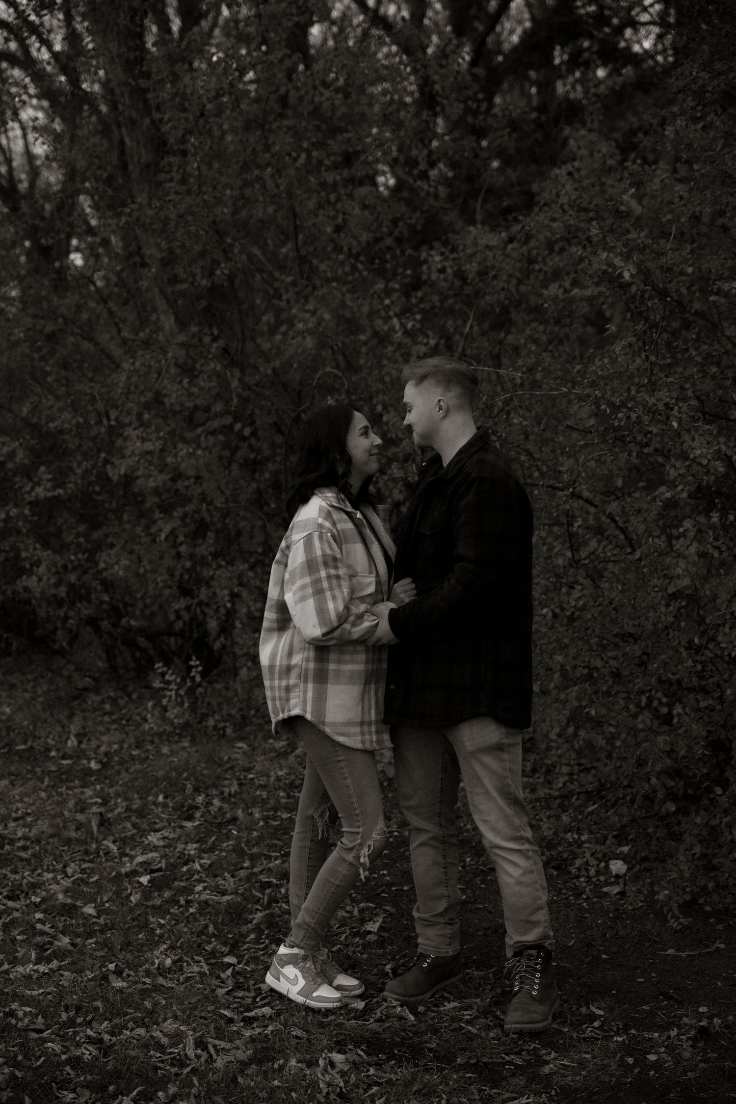 Black and white photo of a couple standing close together in a forest, gazing into each other's eyes romantically.