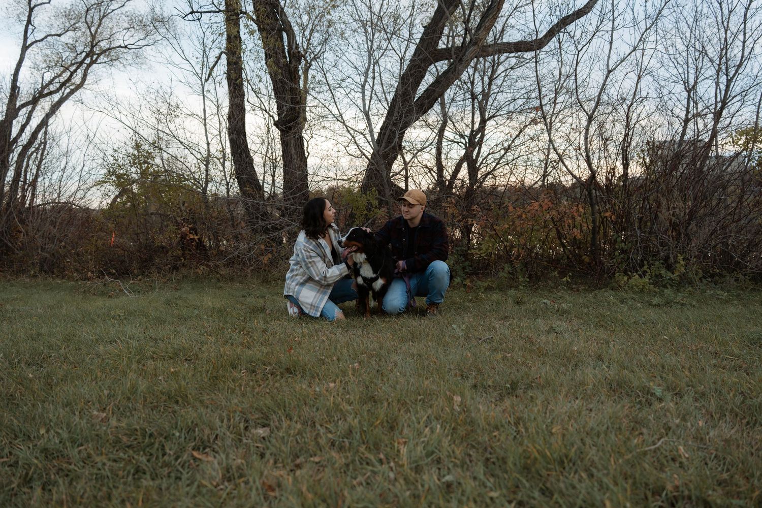 Couple crouching with a small dog in an open grassy field surrounded by bare trees at dusk.