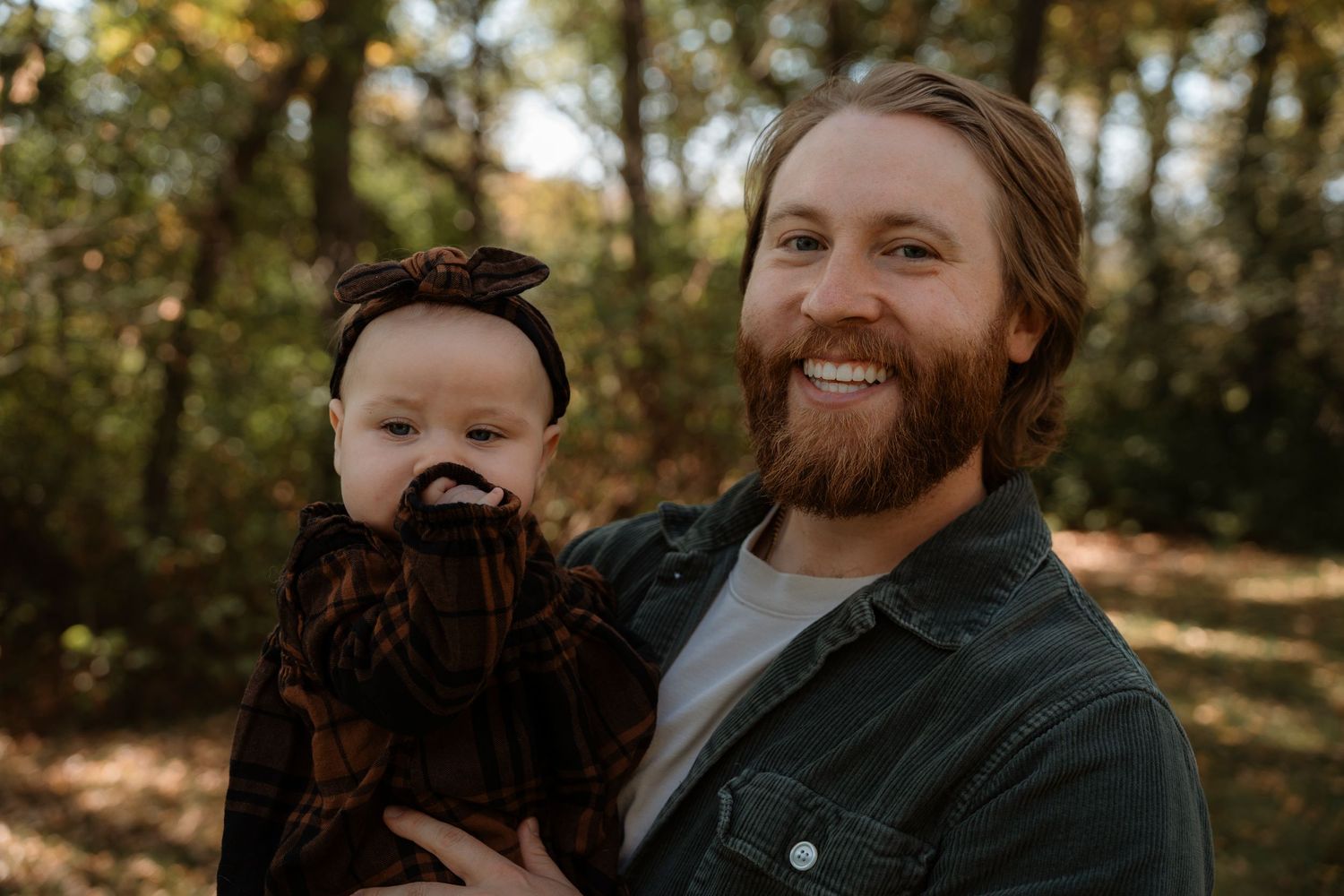 Smiling bearded man holding a baby dressed in dark outfit with bow headband outdoors in autumn forest setting.