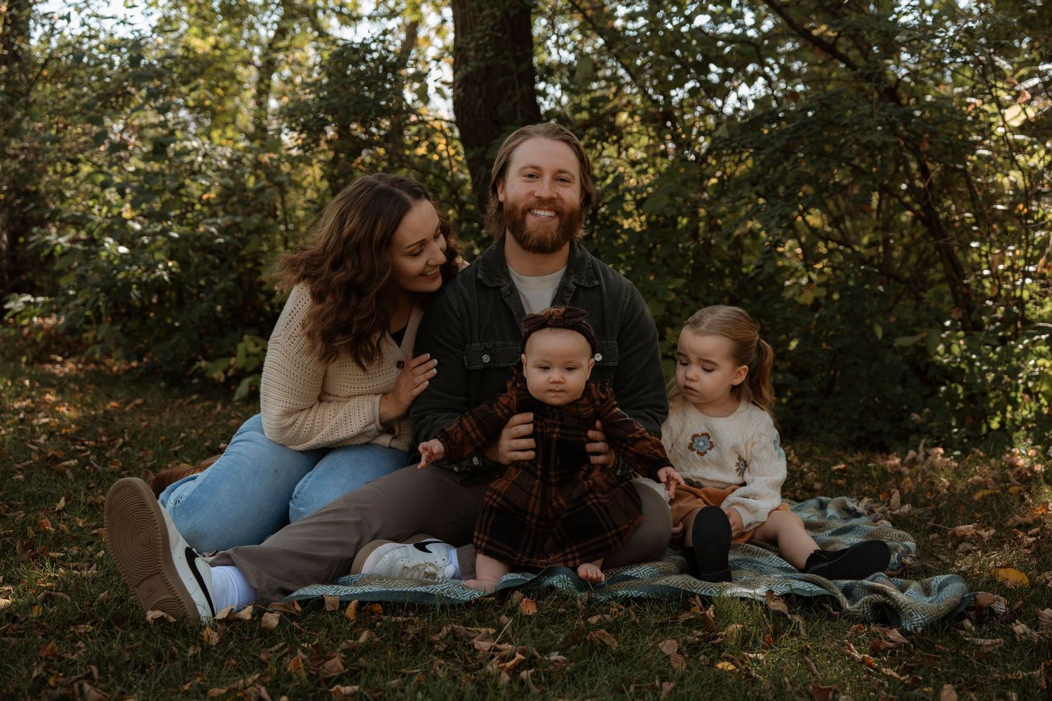 Family of four sitting on a blanket outdoors in a lush green forest, smiling together for a portrait.