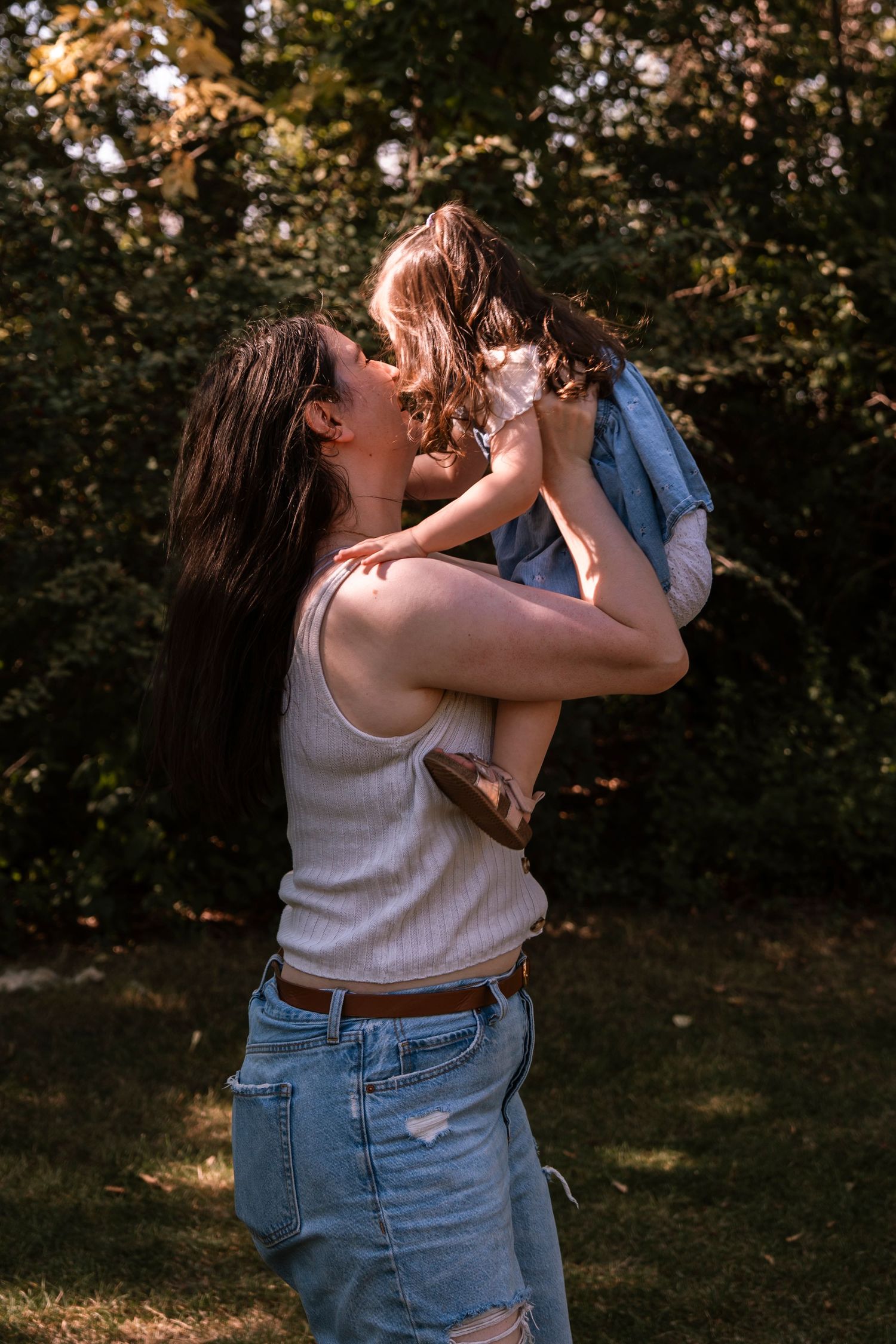Parent in white top and jeans lifts and spins with child in blue shirt while playing outdoors at sunset. Regina Saskatchewan Family portrait