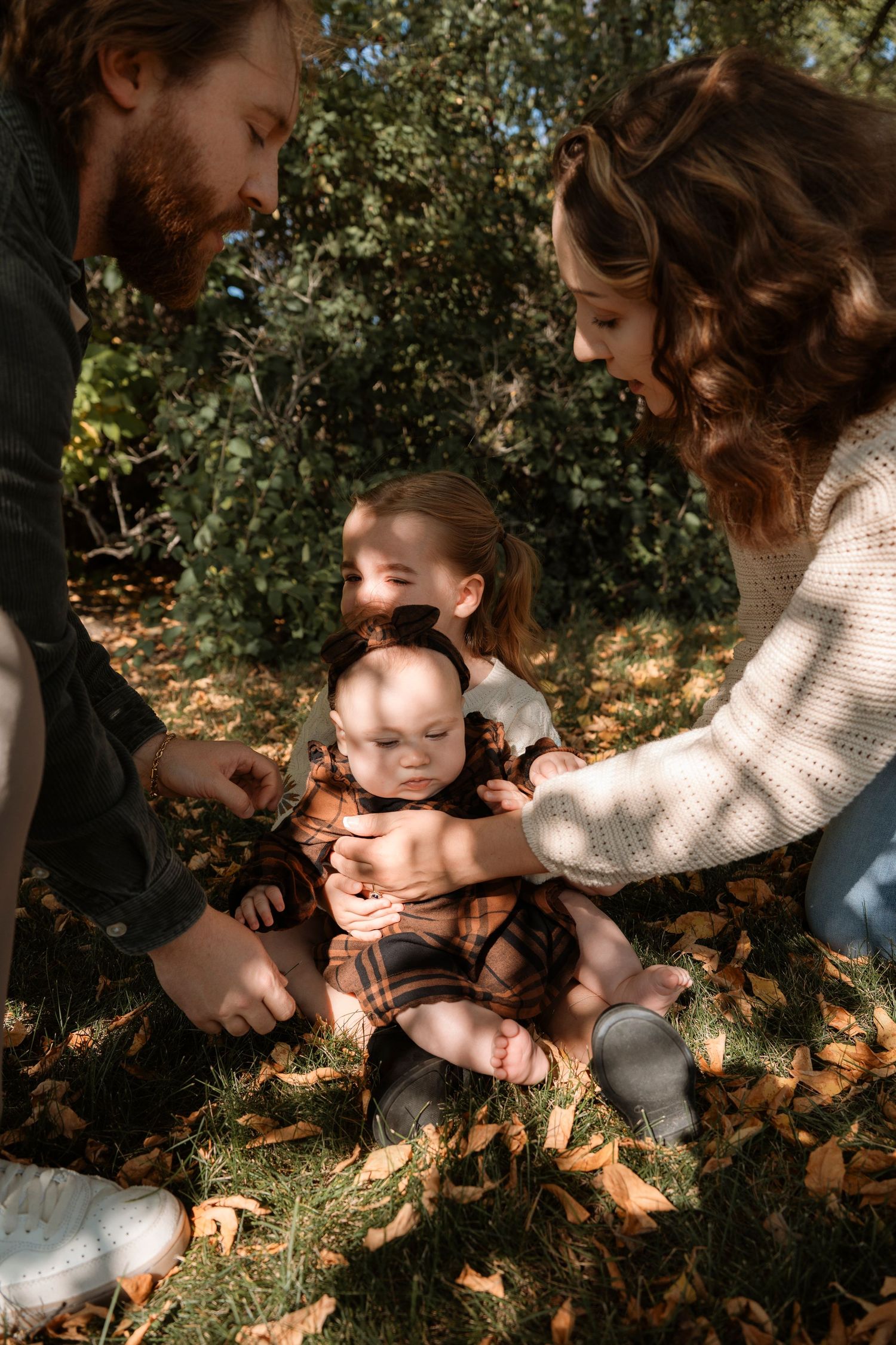 Family with two young children sitting on grass covered in autumn leaves in a sunny park setting.