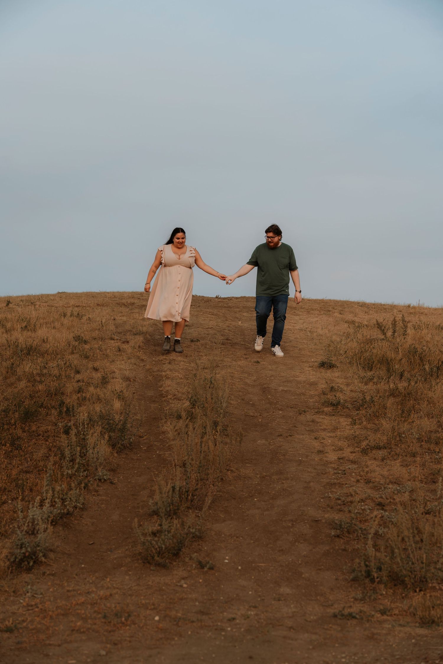 A couple holds hands while walking down a dry grassy hillside, woman in pink dress, man in green shirt and jeans.