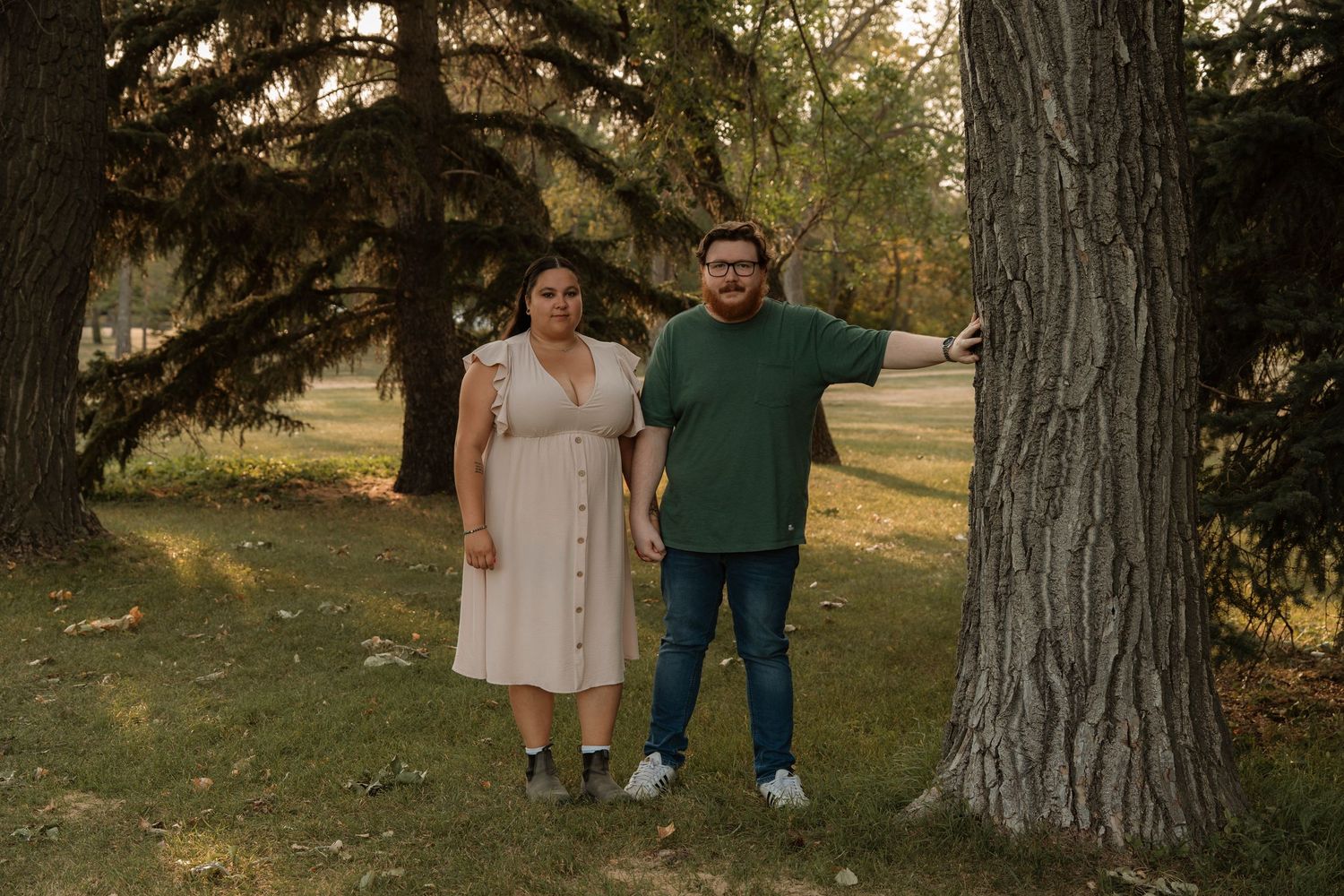 Couple standing together near large trees in a park, woman in beige dress and man in green shirt.