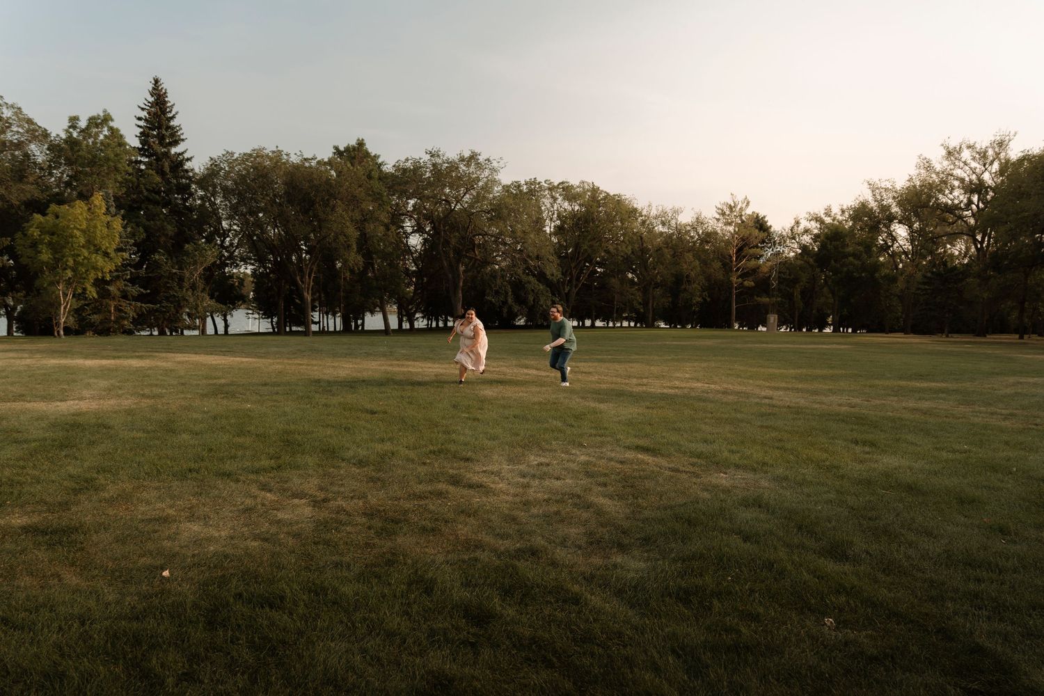 Couple in wedding attire running joyfully across a lush green park lawn surrounded by tall trees at golden hour.