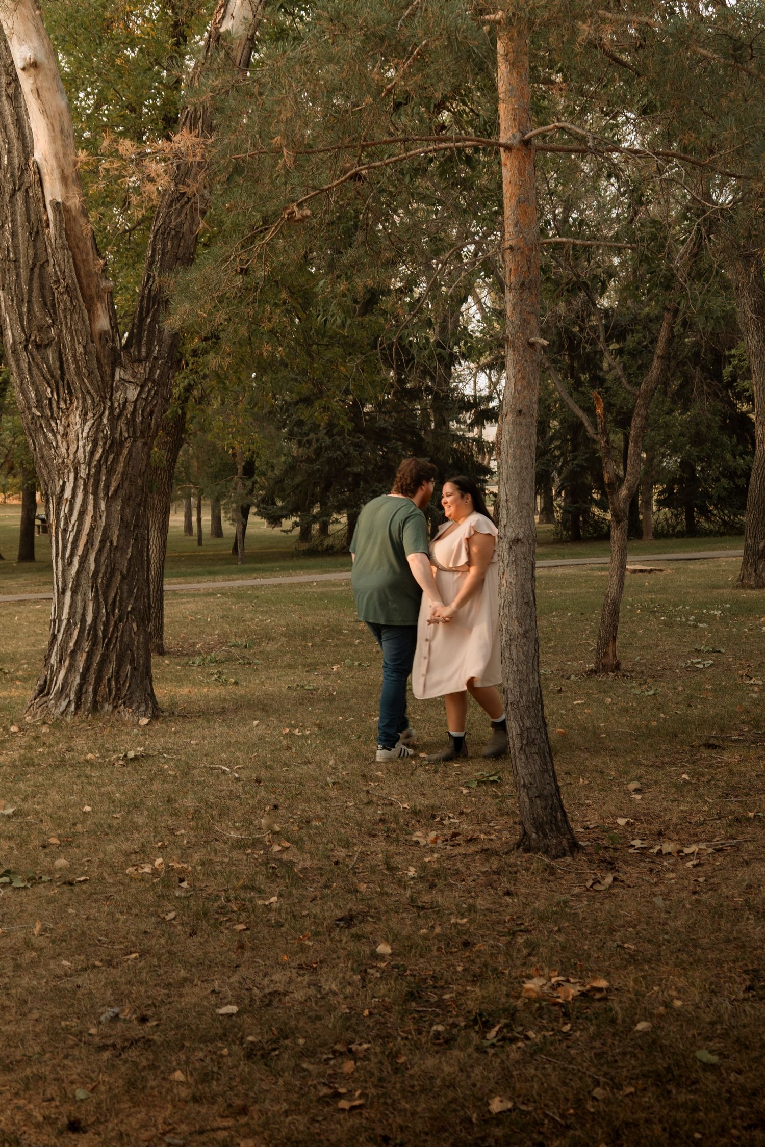 Couple sharing a romantic moment between trees in a lush green park, woman in pink dress holding hands with man in green shirt.