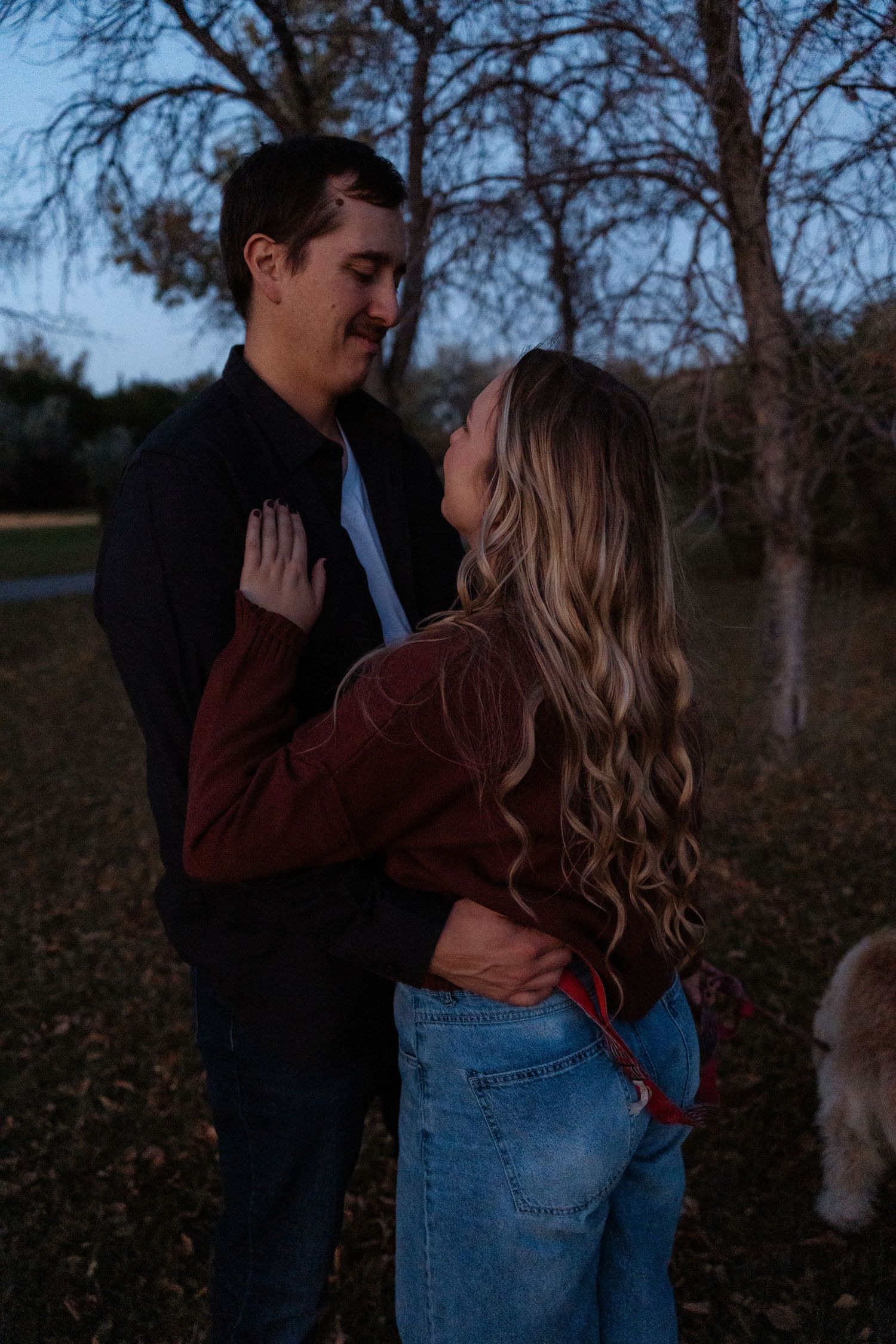 Couple embracing romantically at dusk in a park, woman in jeans and burgundy jacket, man in dark jacket.