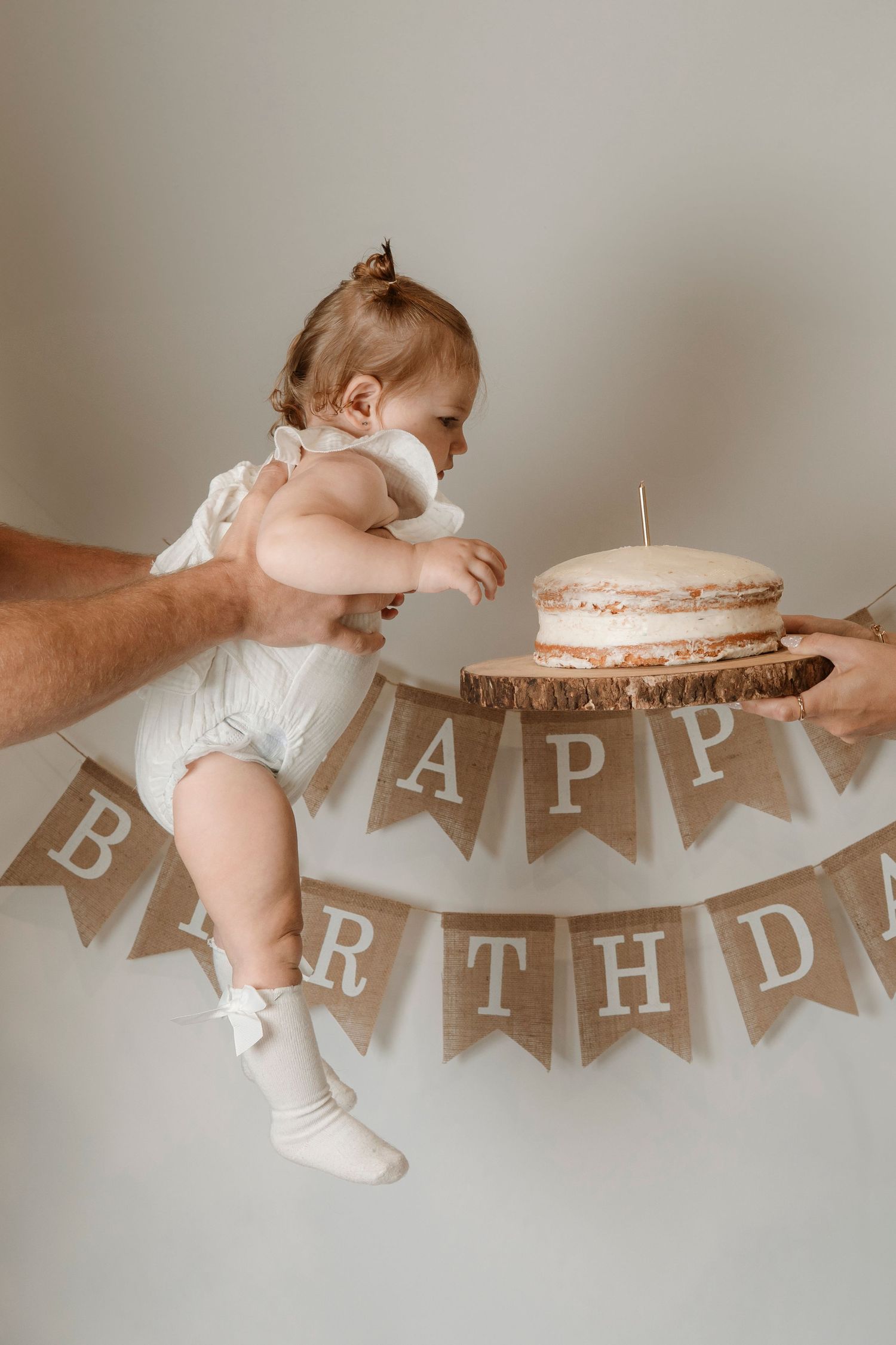 Baby reaching for a rustic naked cake on a wood slice during a first birthday celebration with burlap Happy Birthday banner.