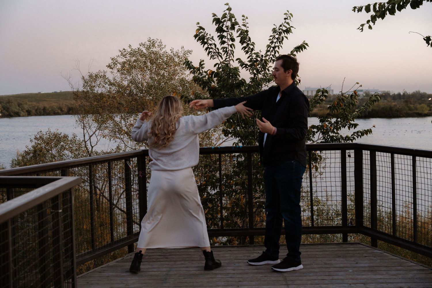 Couple dancing together on a wooden deck overlooking a serene river at dusk, surrounded by autumn trees.