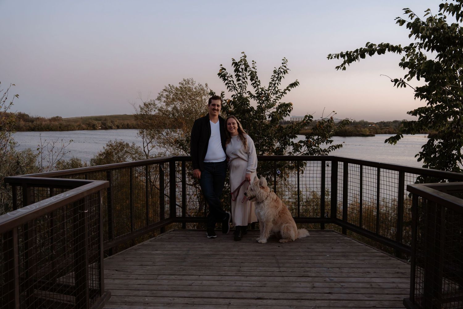 Couple posing with golden dog on wooden deck overlooking scenic lake at dusk with autumn trees.