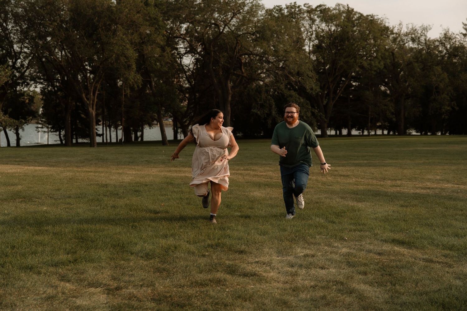 A joyful couple runs through a lush green park at golden hour, surrounded by tall trees near a lake.
